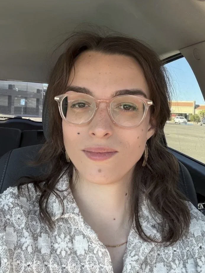 A young woman with glasses and shoulder-length wavy brown hair sitting in a car, wearing a patterned shirt, with a view of a parking lot and stores outside.