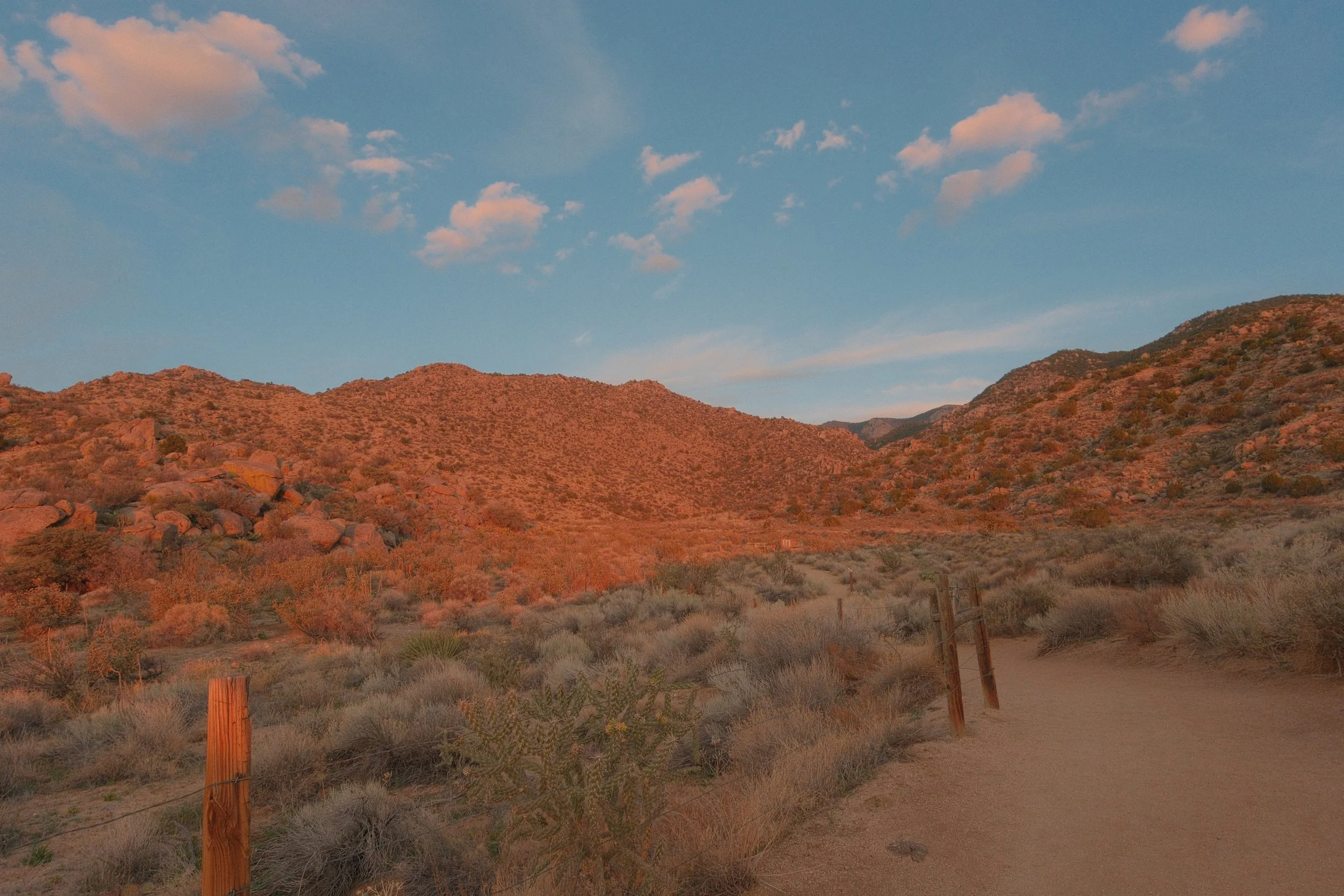 A desert landscape with reddish-brown hills, a dirt trail, and wooden posts along the path under a blue sky with scattered pink clouds.