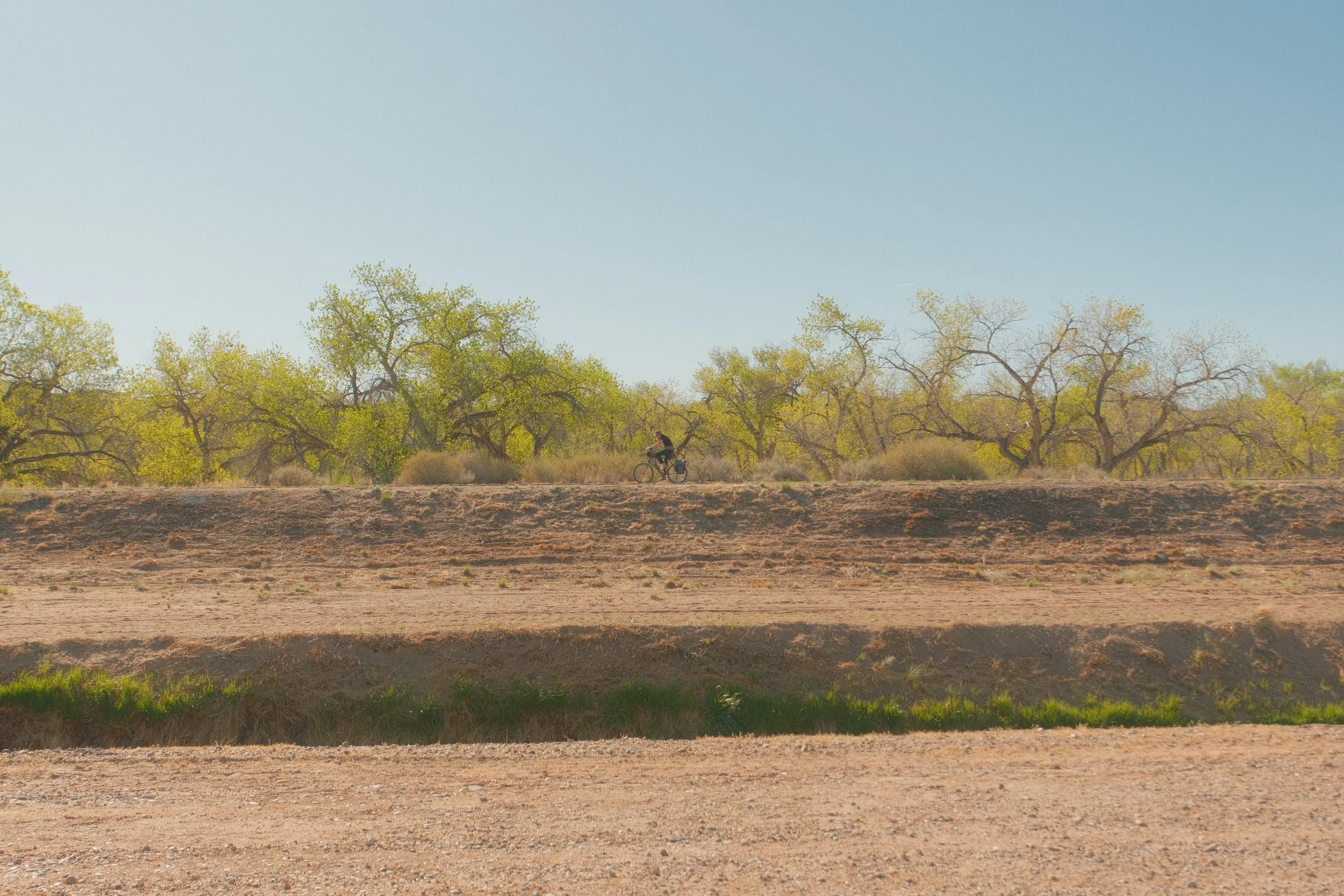 A person riding a bicycle along a dirt path in a semi-arid landscape with sparse green trees and bushes under a clear blue sky.