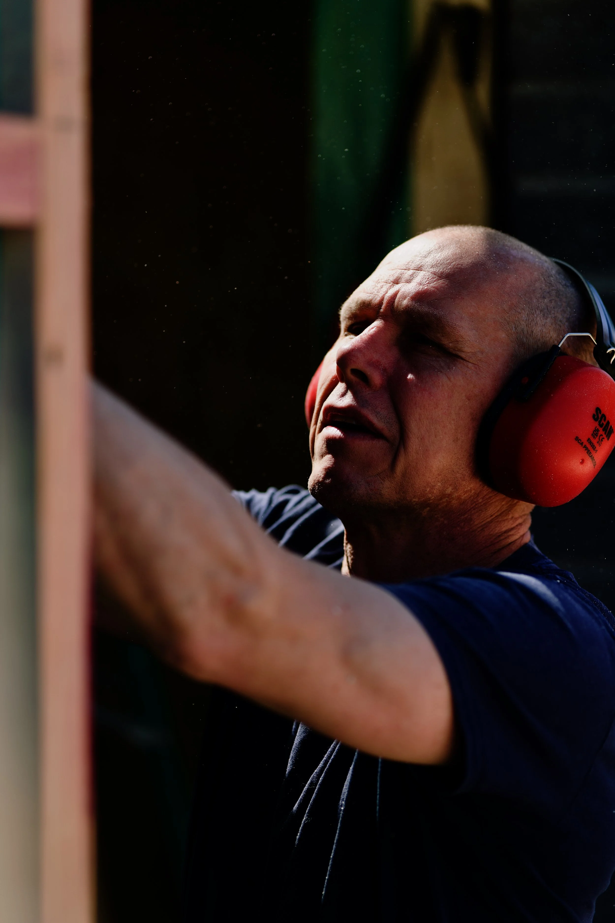 Mark wearing red ear protection and a Ralphs Building Services shirt. Concentrating on an activity, possibly woodworking or construction, with a blurred background.