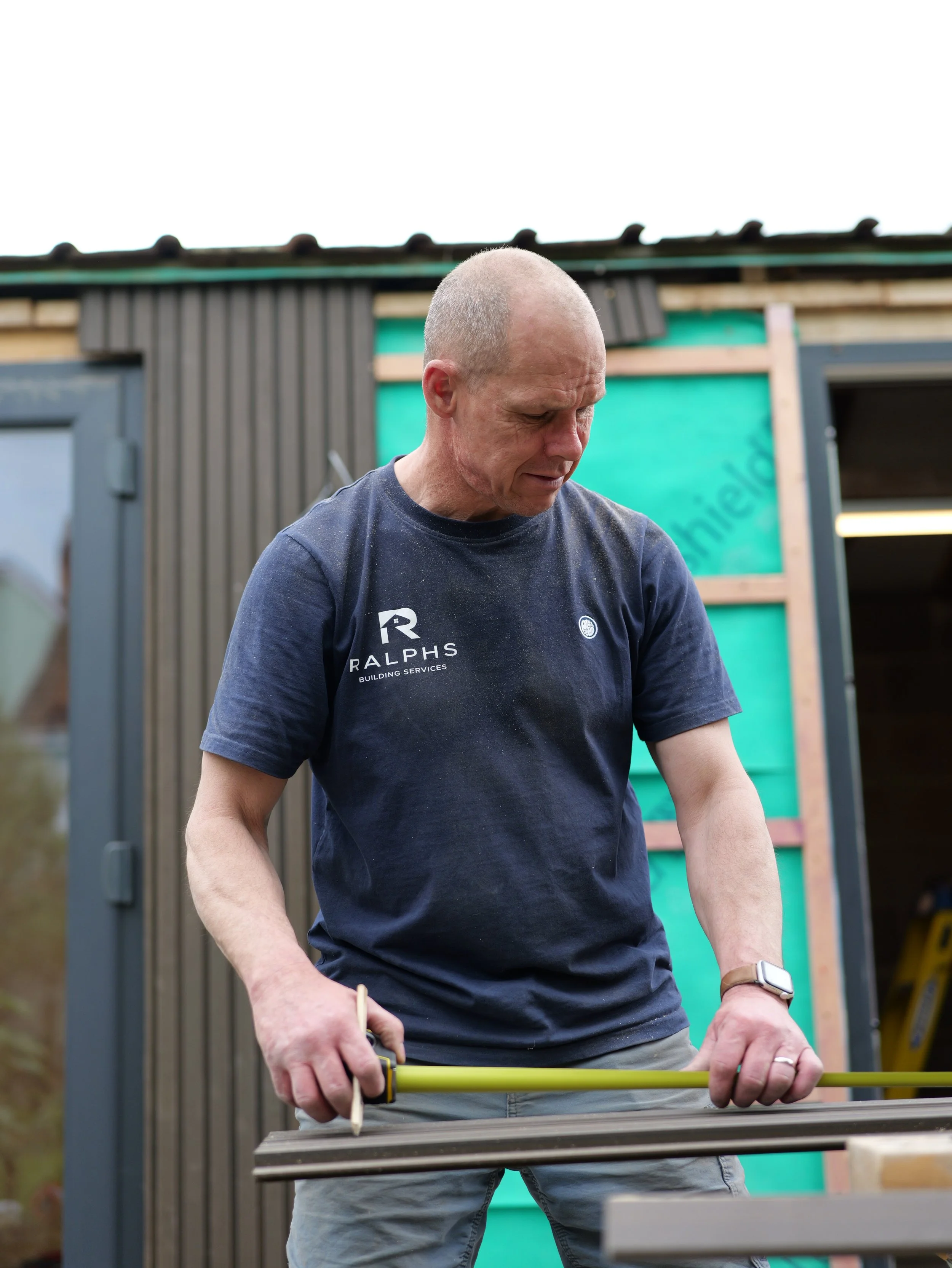 Mark Ralphs, builder, measuring timber on site wearing a Ralphs Building Services t-shirt