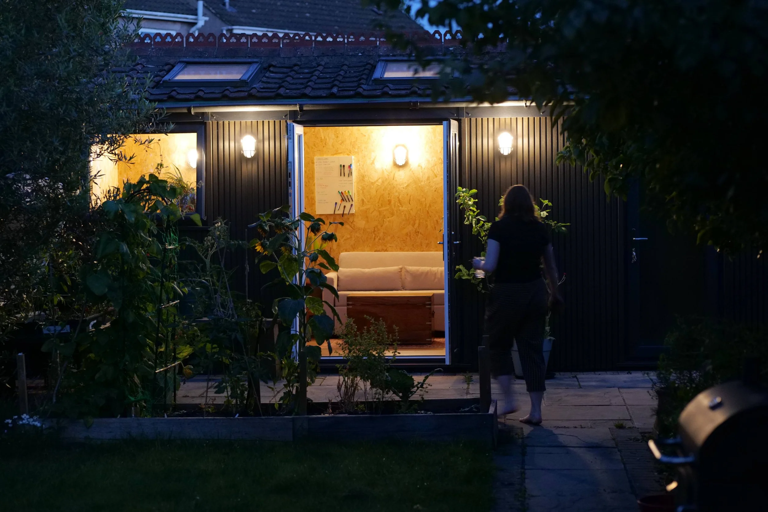 Garden room exterior at dusk, illuminated from within, with a person approaching the open doors