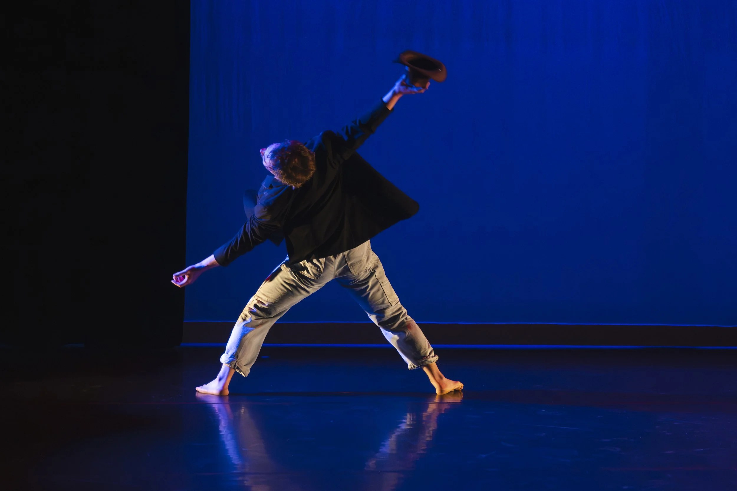 A person performing a dance move on a dark stage illuminated by blue lighting, holding a hat in one hand and balancing on one foot with the other leg extended.