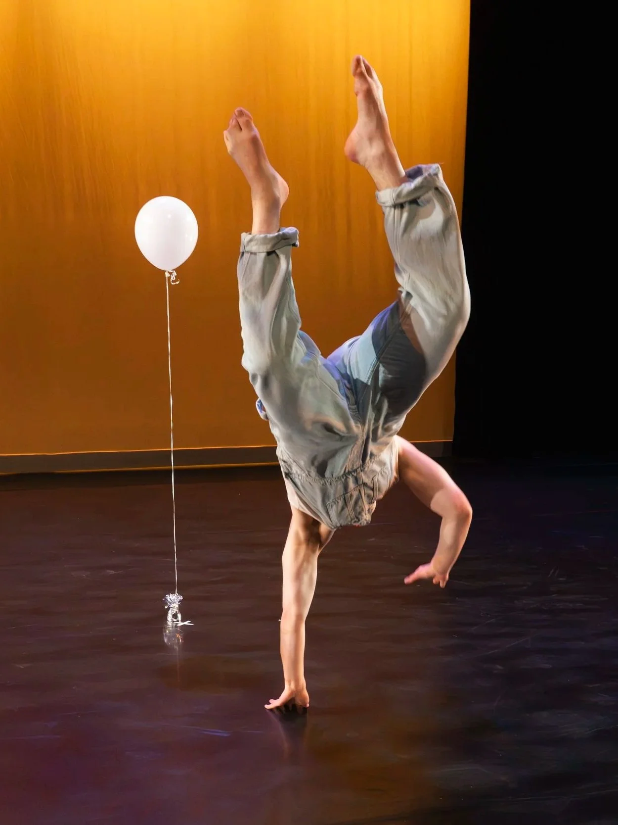 A person performing a handstand on one hand, with their legs extended upwards, in a theater. A white balloon tied to a string is on the ground nearby.