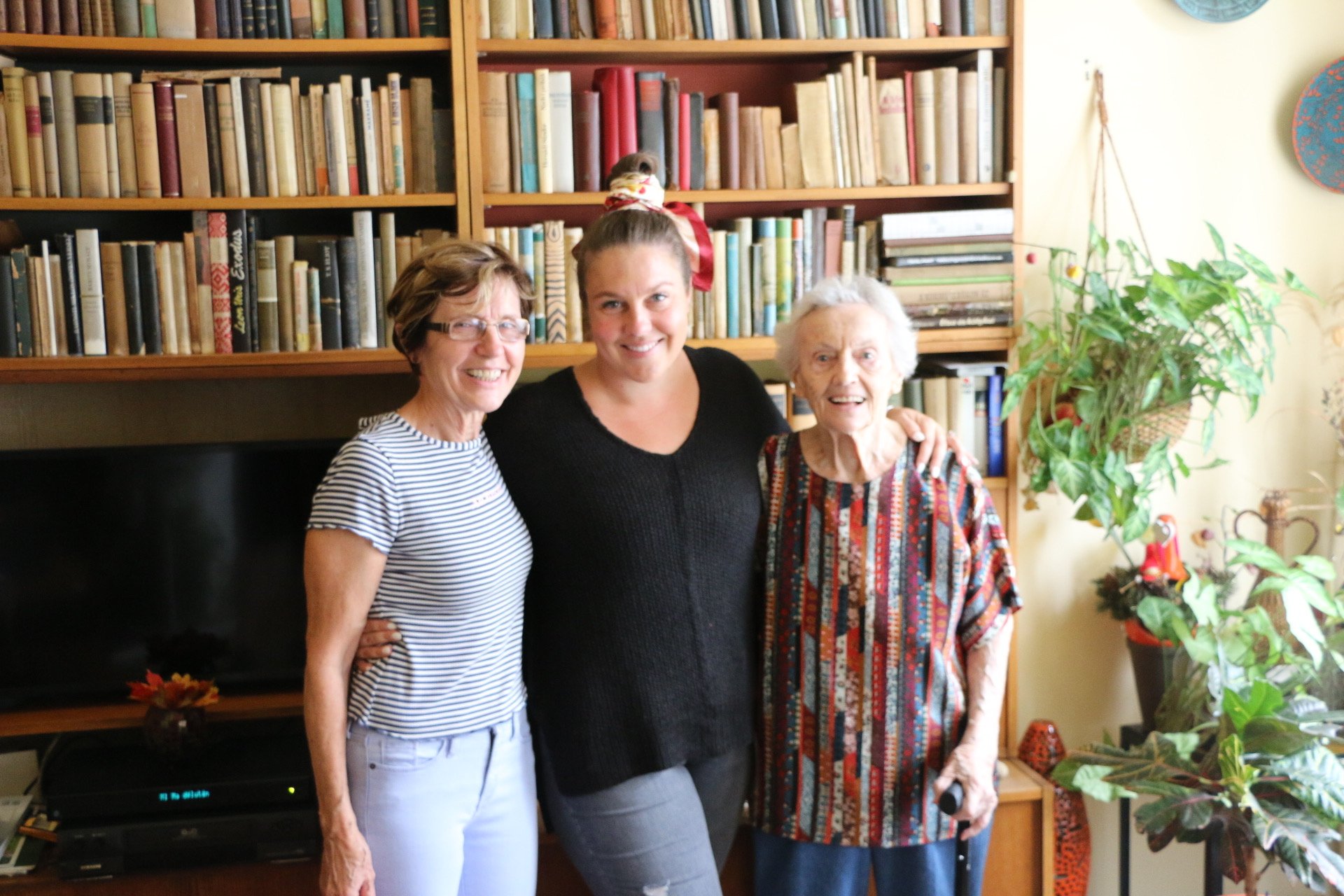 Three smiling women posing together in front of a large bookshelf in a cozy living room.