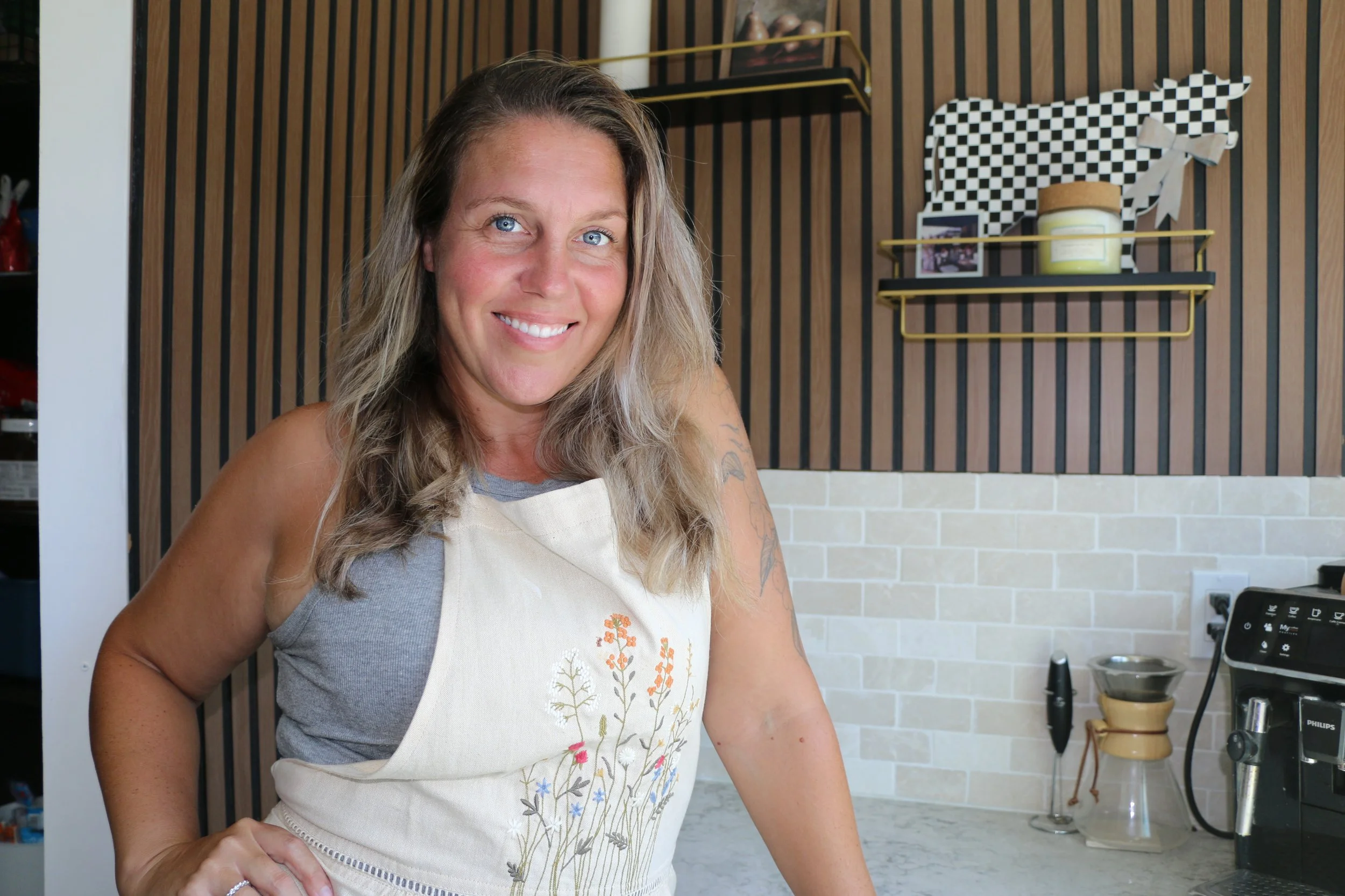 Smiling woman wearing a floral apron in a modern kitchen with a coffee maker.