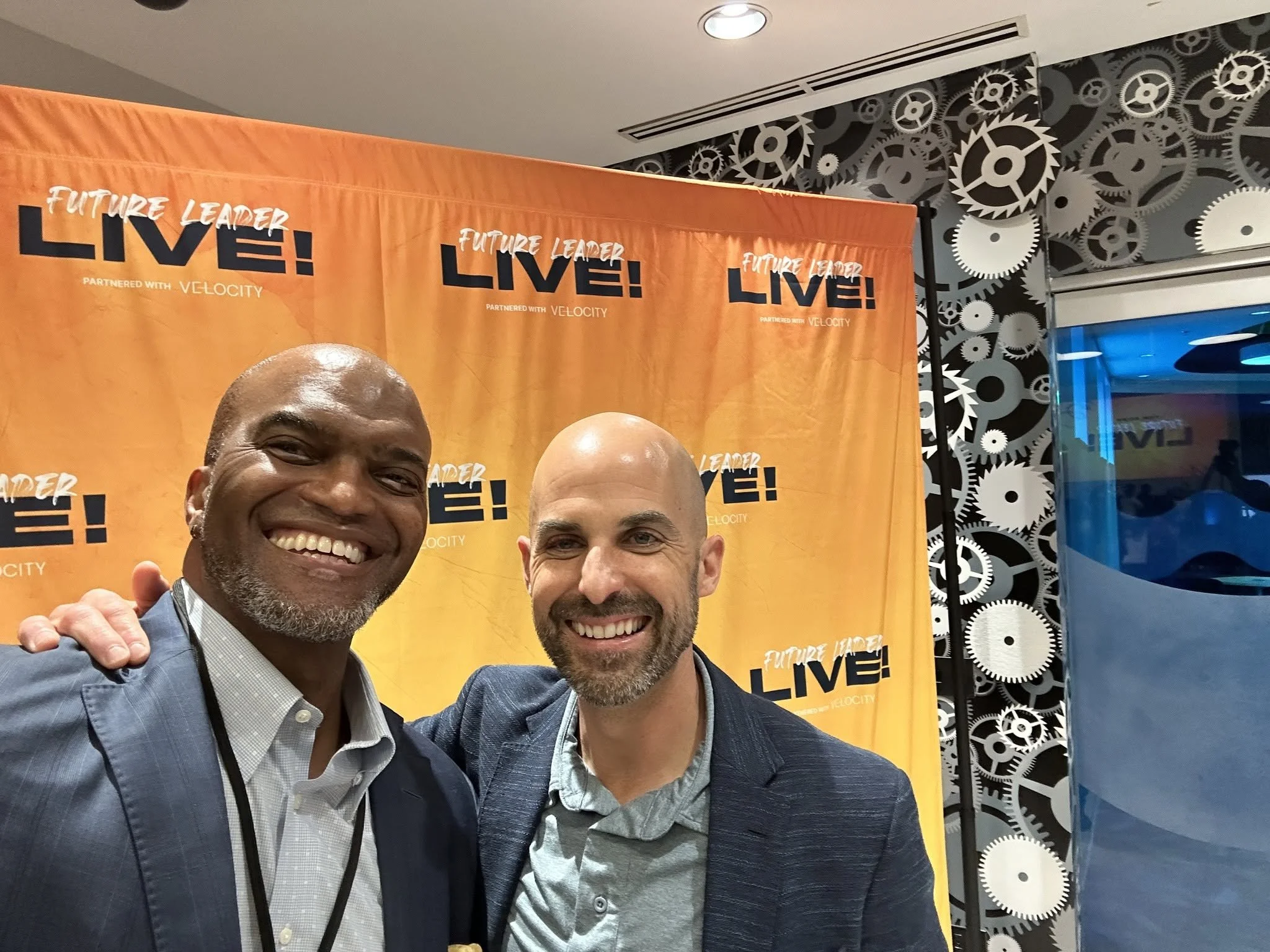 Two men, Toby Brazwell and Mark Ostach, smiling at the camera in front of an orange and black backdrop with the words "Future Leader LIVE!" and "Partnered with Velocity" and a background of gears, in an indoor setting.