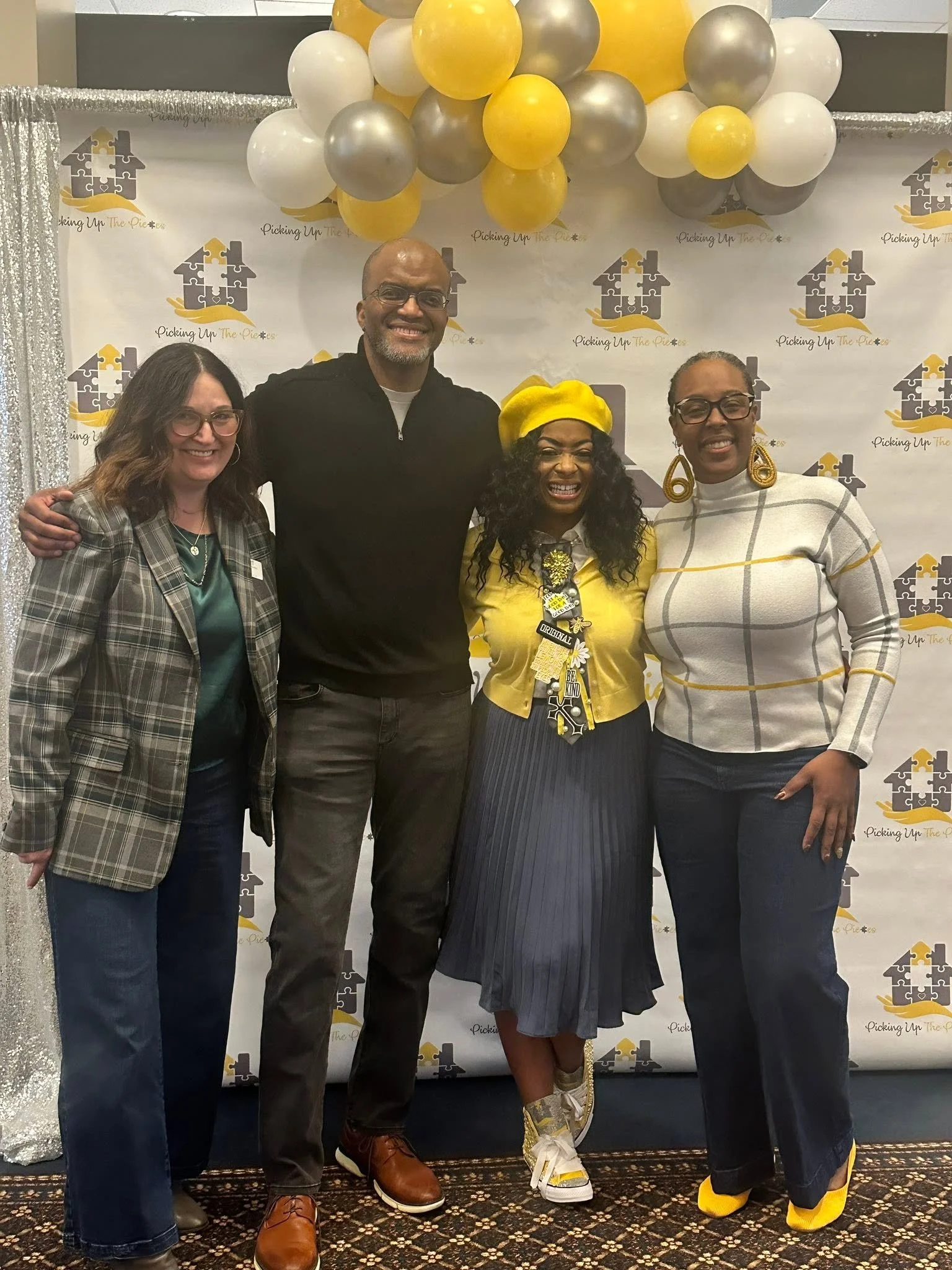 Four people, including Toby Brazwell, standing together at an event with a backdrop that says 'Picking Up The Pieces' and decorated with yellow, white, and gray balloons. They are smiling and dressed in casual to semi-formal attire.