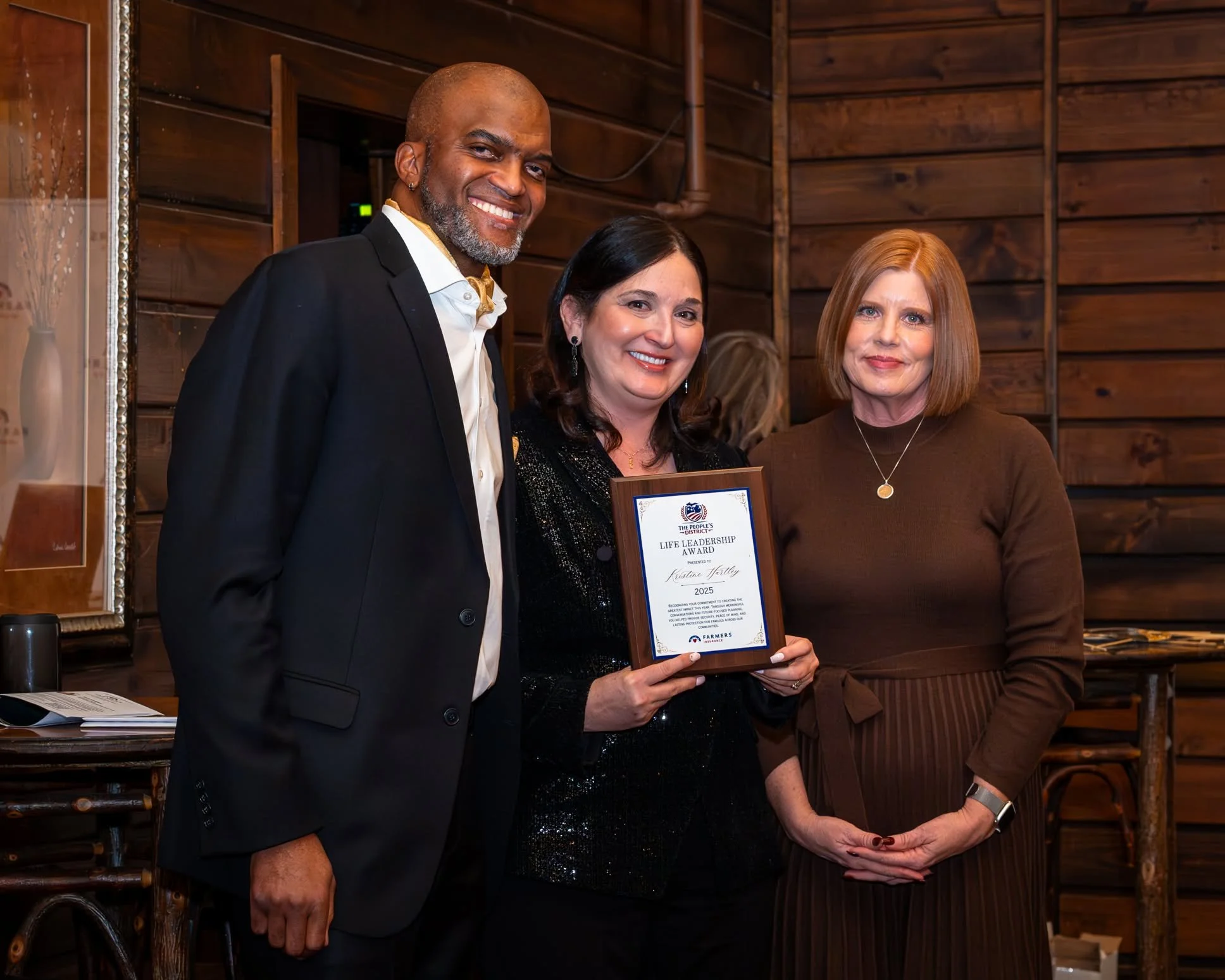 Three people standing together indoors,  with Toby Brazwell and a woman holding a framed award. The man is smiling, the woman is also smiling, and the other woman has a neutral expression. The background features wooden paneling.