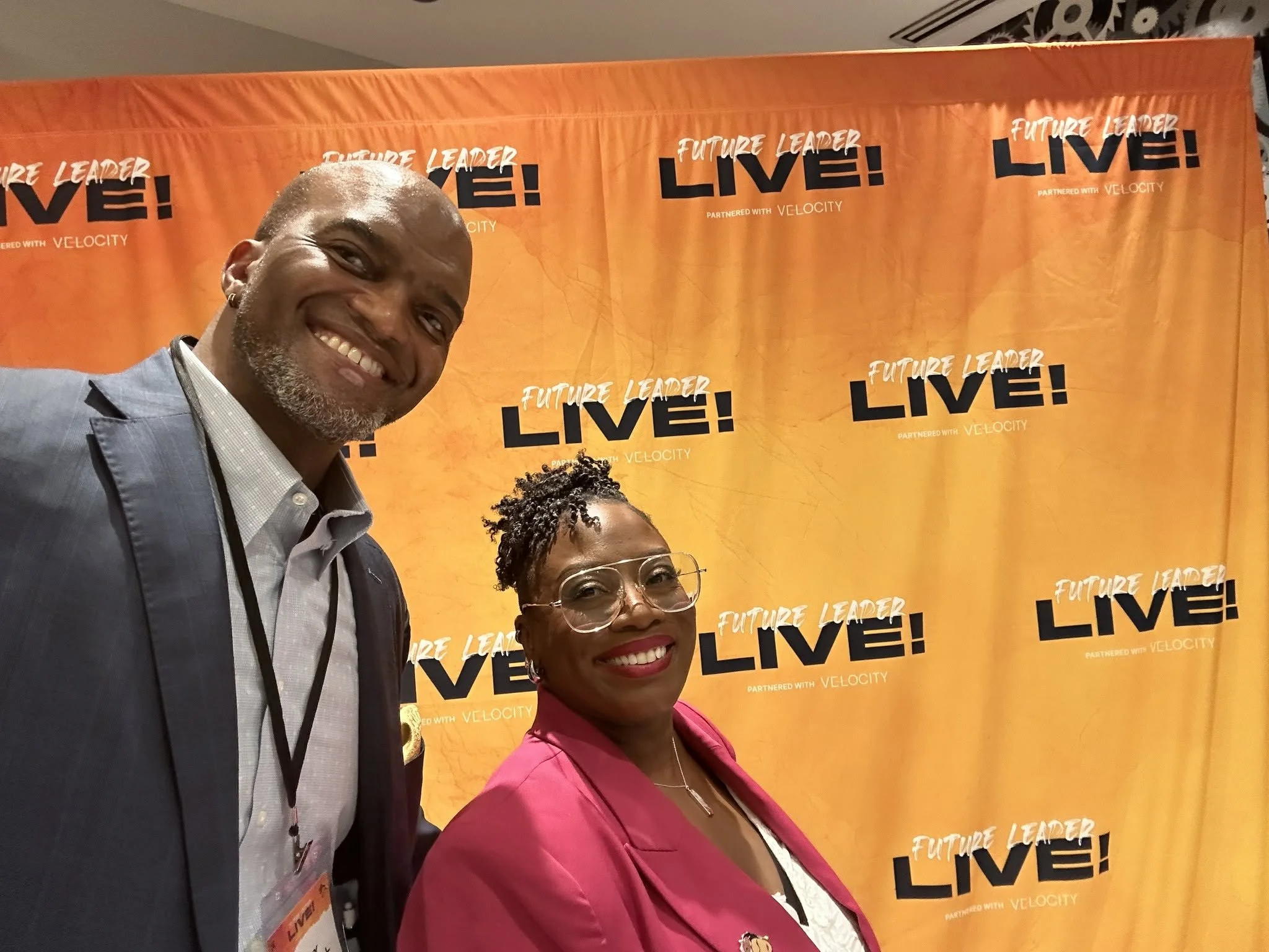 Smiling Toby Brazwell and a woman standing in front of an orange backdrop with the words "Future Leader Live! Partnered with Velocity" repeated multiple times.