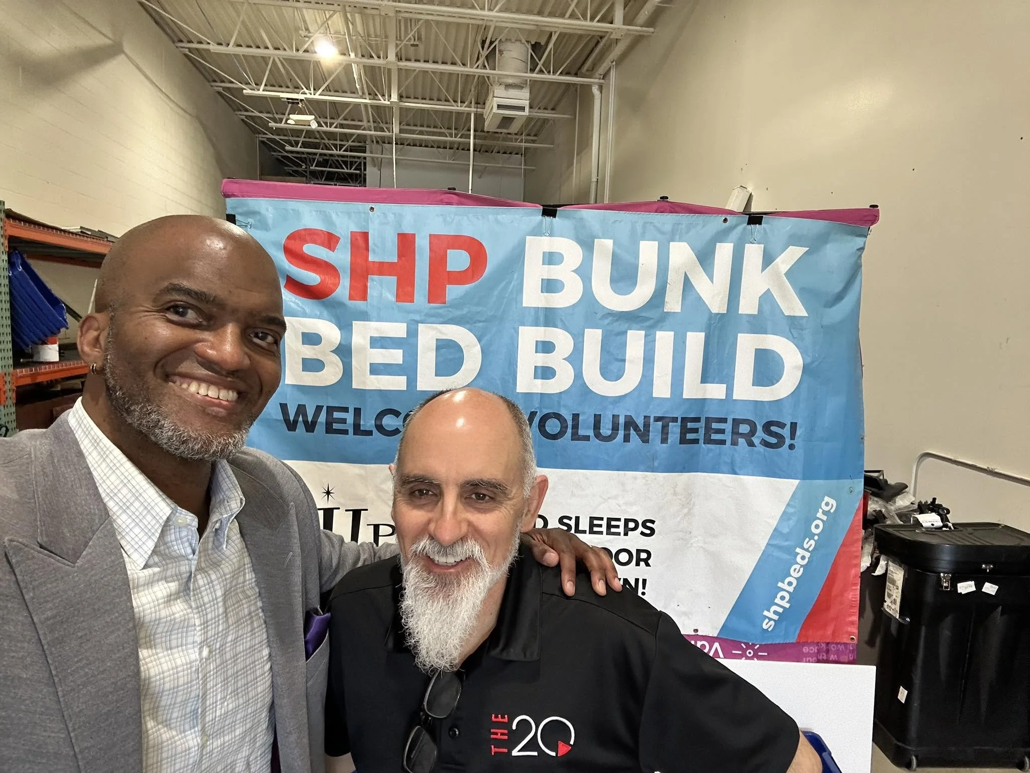 Two men smiling and posing for a photo in front of a blue banner that reads 'Ship Bunk Bed Build, Welcome Volunteers'. The man on the left is bald with a gray beard, wearing a gray suit jacket and a white shirt. The man on the right has a bald head and a white beard, wearing a black t-shirt with red text. The setting appears to be an indoor warehouse or workshop with shelves and equipment in the background.