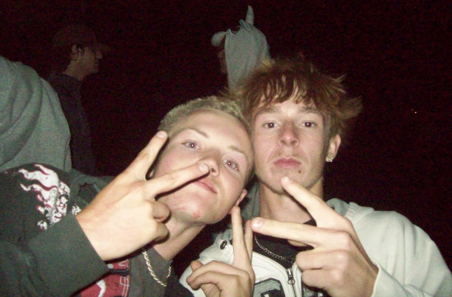 Two young men taking a selfie at night, both making peace signs with their hands.