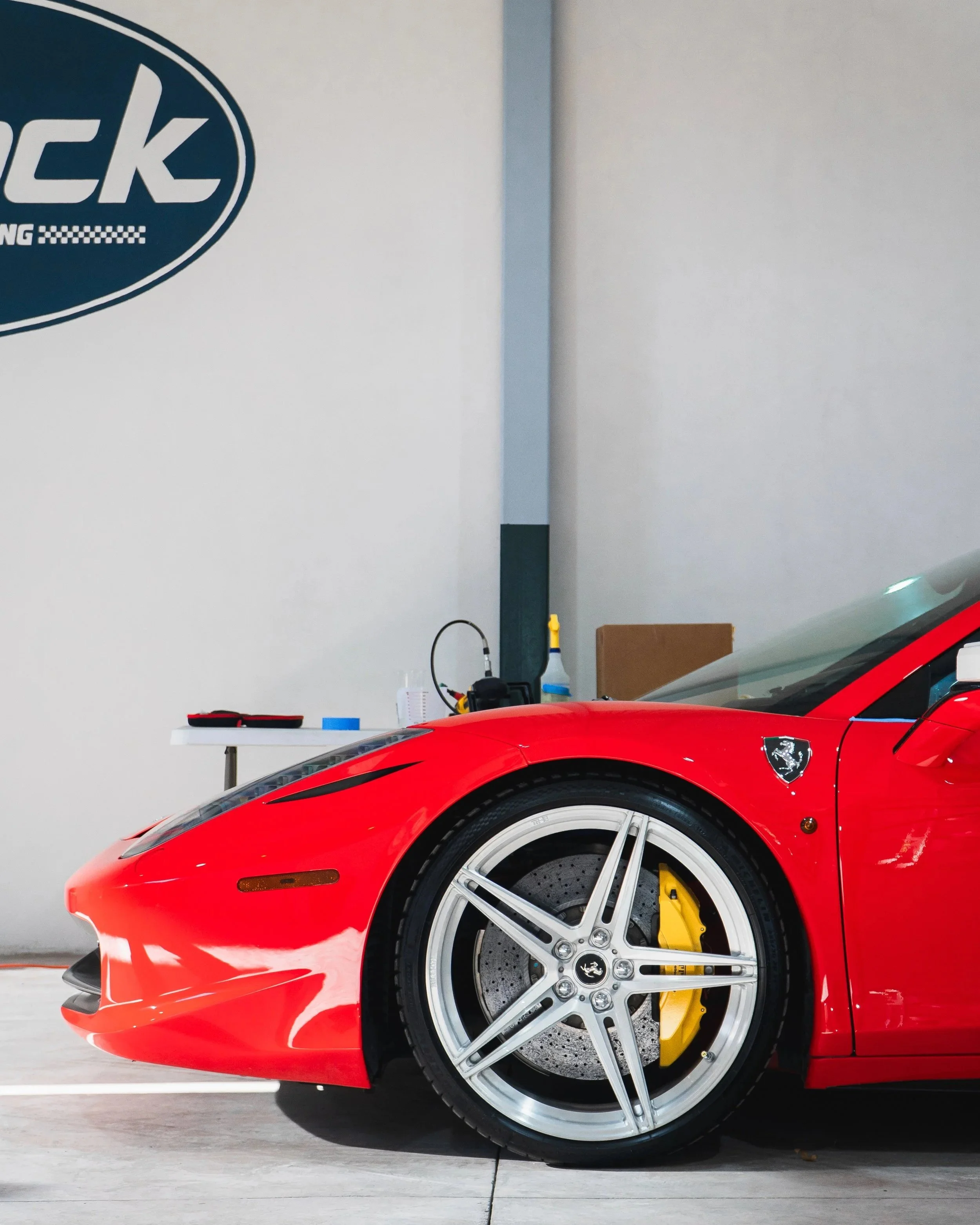 El lado frontal de un coche deportivo rojo, específicamente un Ferrari, con neumático grande, frenos amarillos y detalles en negro, en un espacio interior con pared blanca y equipo de trabajo en el fondo.
