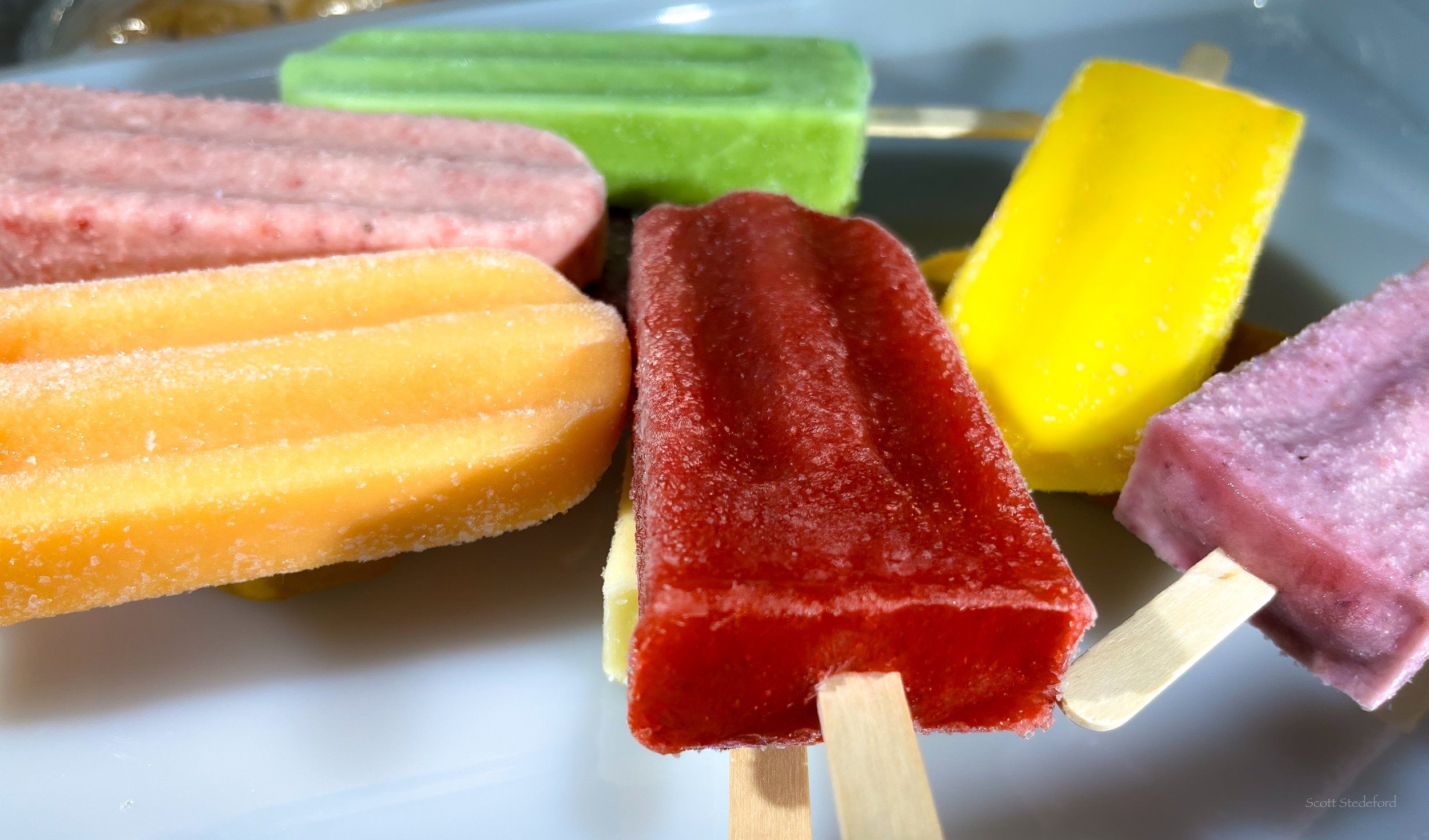 Close-up of colorful frozen fruit popsicles with various flavors including yellow, red, pink, green, and orange on a white surface.