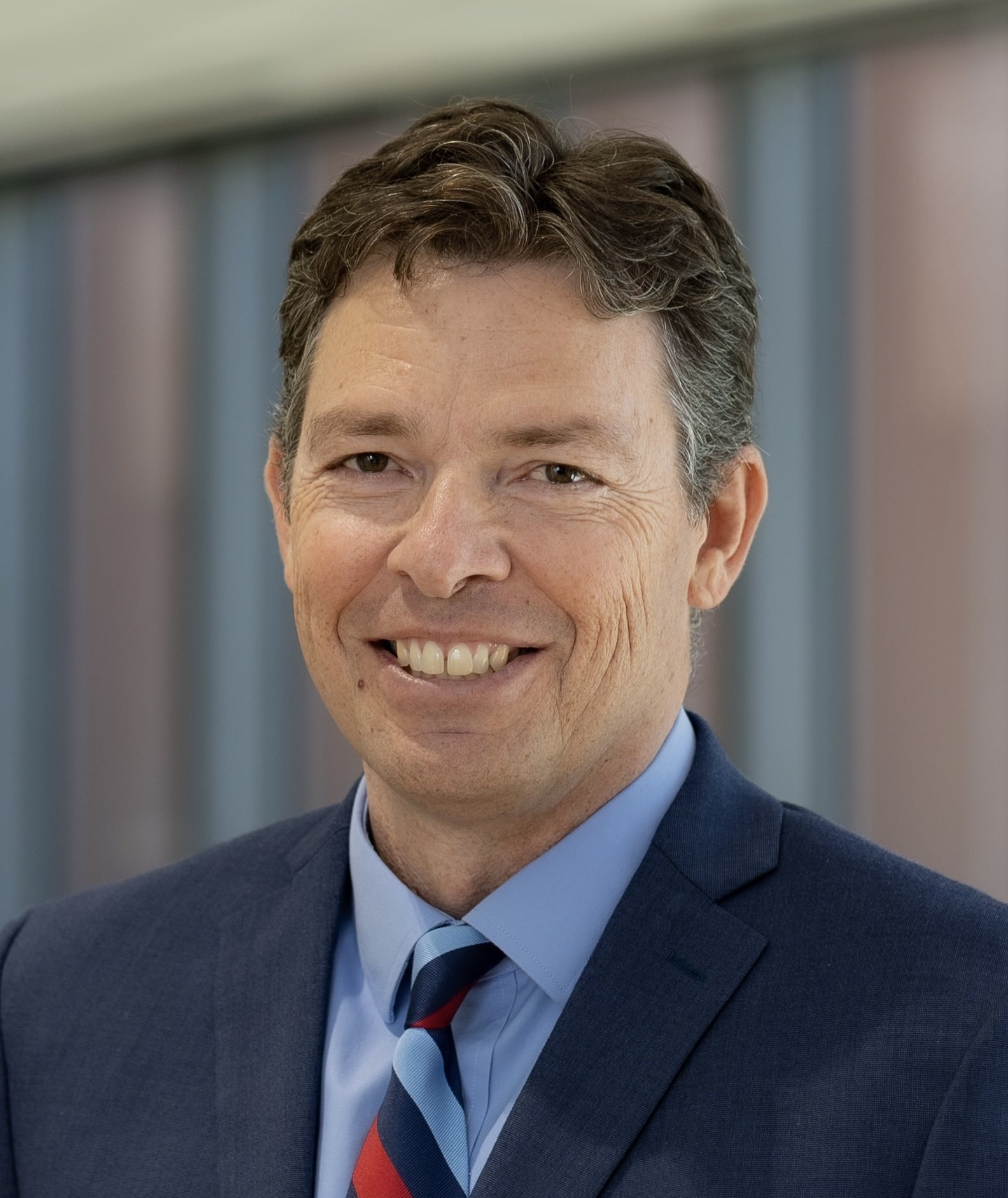 Close-up photo of a smiling middle-aged man with dark brown hair and light skin wearing a dark blue suit, light blue shirt, and a striped blue and red tie, standing in front of a blurred background of what looks like a building or office environment.
