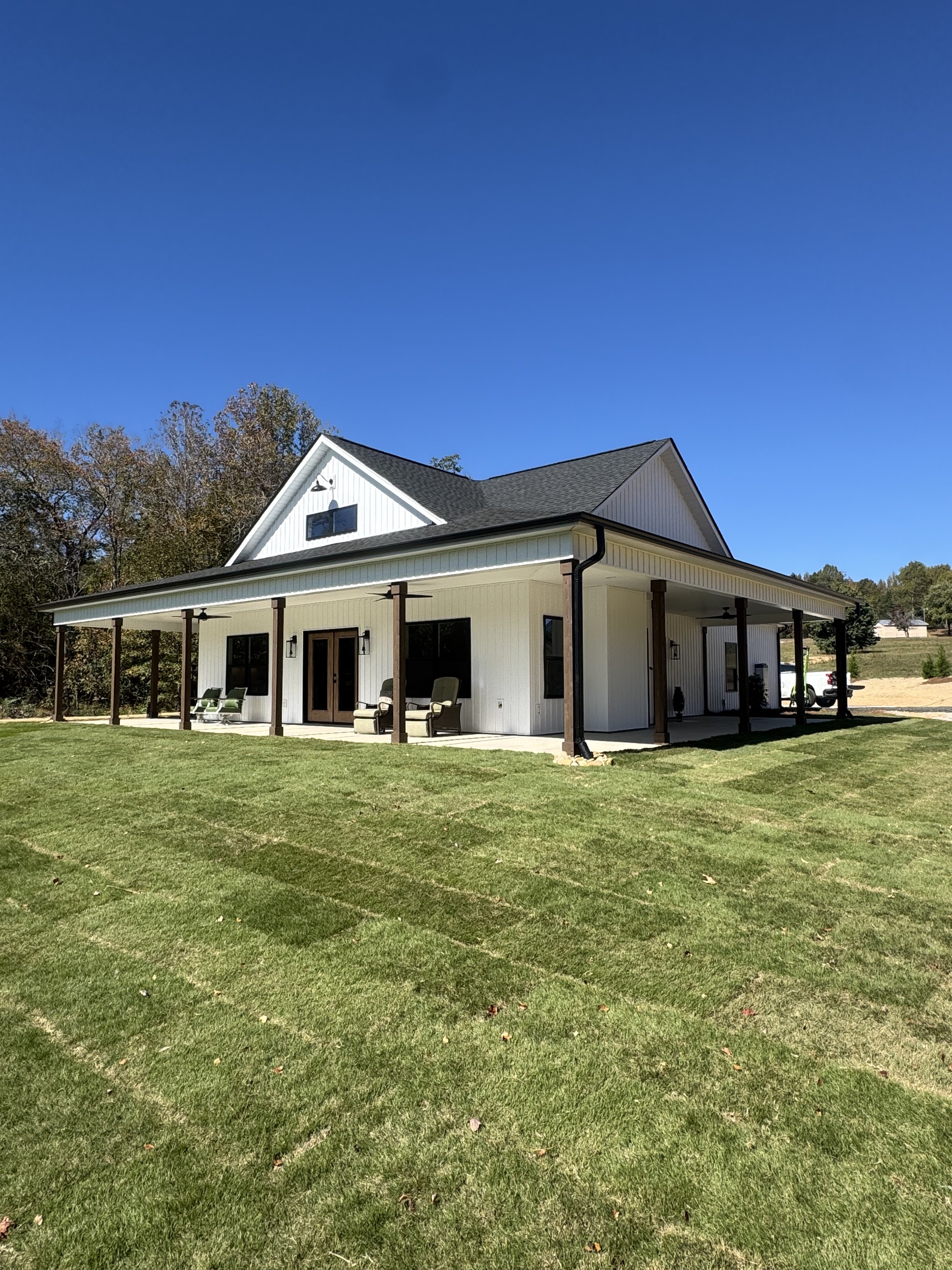 A white house with black roof and trim features a covered porch with outdoor furniture, set on a well-manicured lawn under a clear blue sky with some trees in the background.