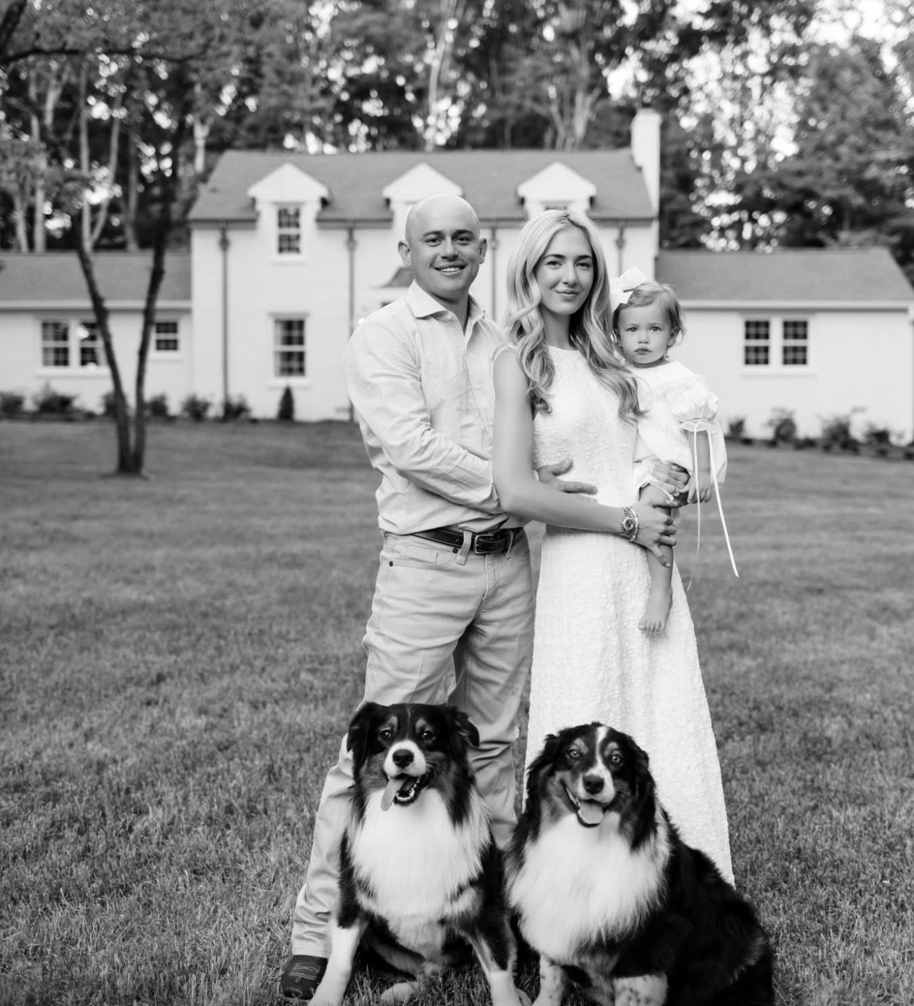 A black and white photo of a family of three standing outdoors in front of a large house with trees in the background. The man is on the left, bald, wearing a light-colored shirt and pants. The woman is in the middle, with long wavy hair, wearing a sleeveless dress, holding a young girl with a bow in her hair. The family is accompanied by two Australian Shepherd dogs sitting in front of them, all on a grassy lawn.