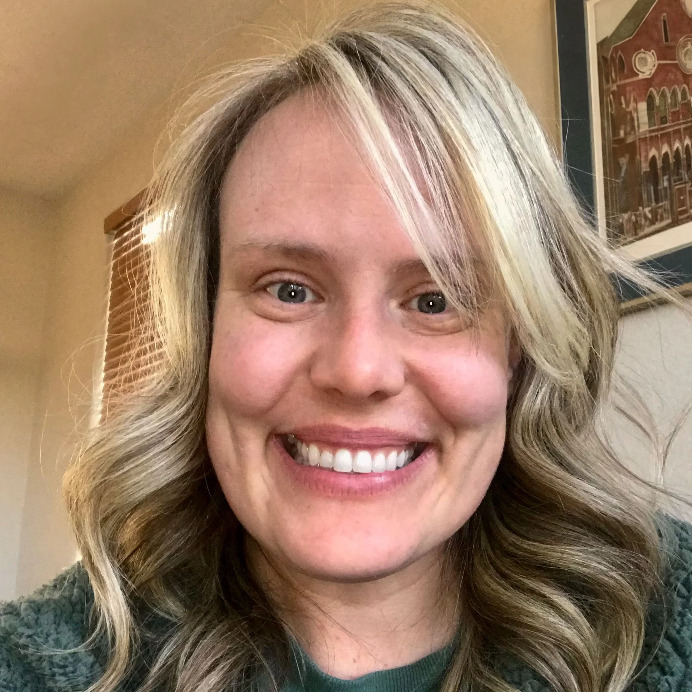 Close-up of a smiling woman with blonde, wavy hair and blue eyes, wearing a green top.