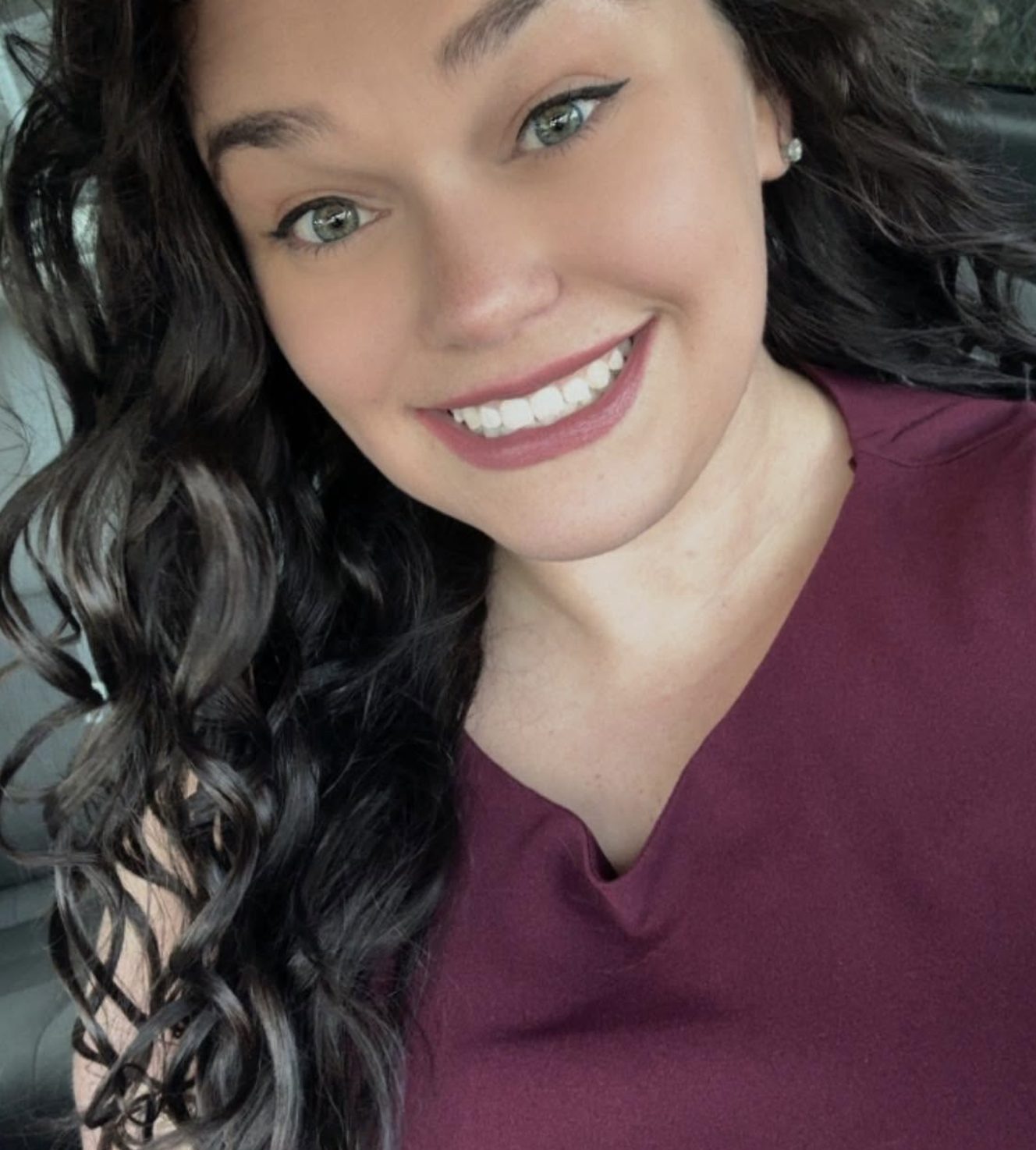 Close-up photo of a woman with long curly dark hair, light skin, and blue eyes, smiling and wearing a maroon shirt with earrings.