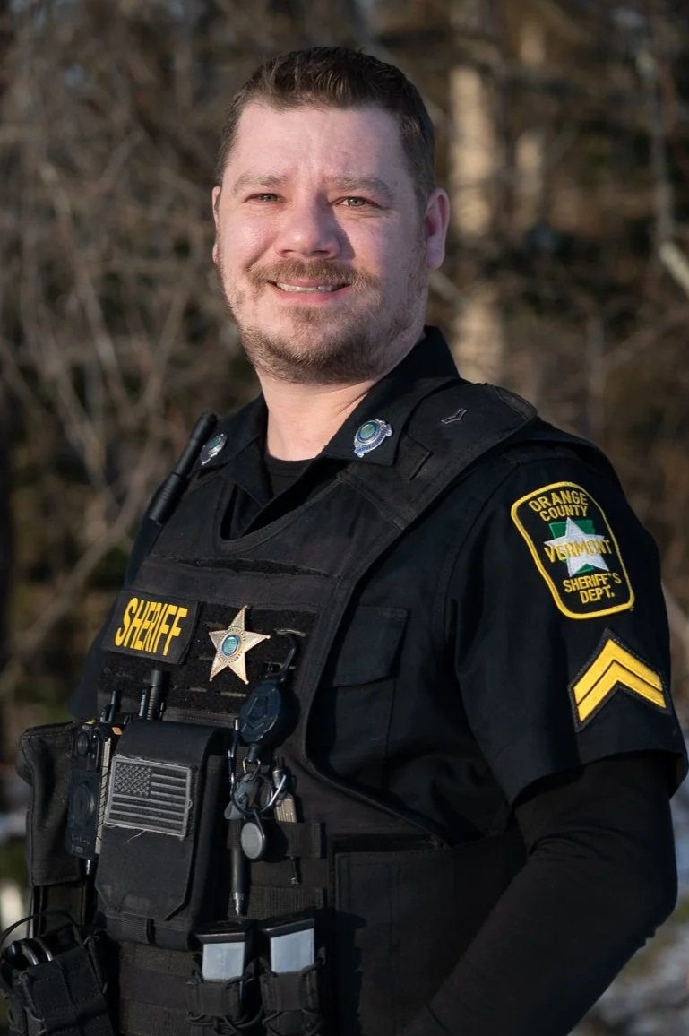 A smiling male sheriff in uniform, standing outdoors with trees in the background.