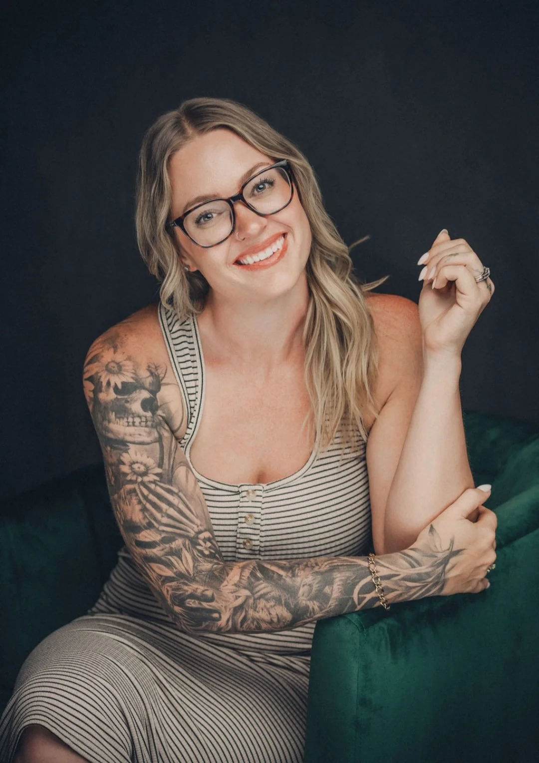 A smiling woman with long wavy blonde hair, glasses, and tattoos on her right arm, wearing a striped tank top and sitting next to a green piece of furniture.