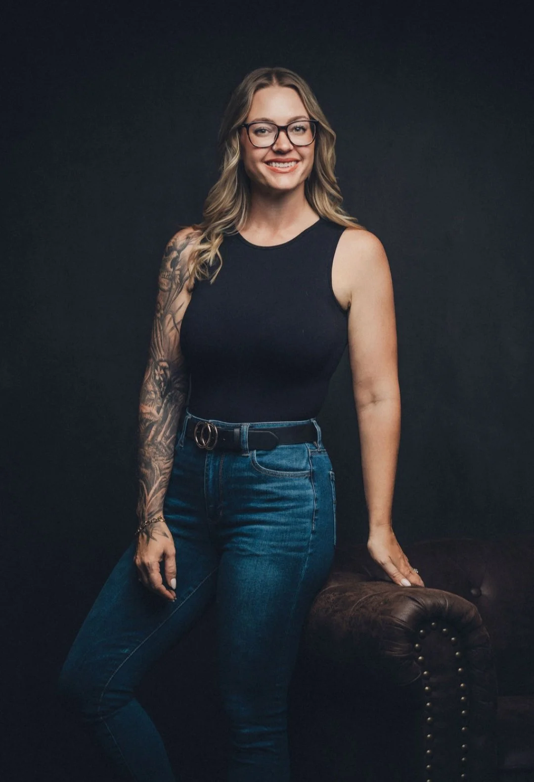 A woman with glasses and tattoos smiling, wearing a sleeveless black top and blue jeans, standing against a dark background and leaning on a dark brown leather chair.
