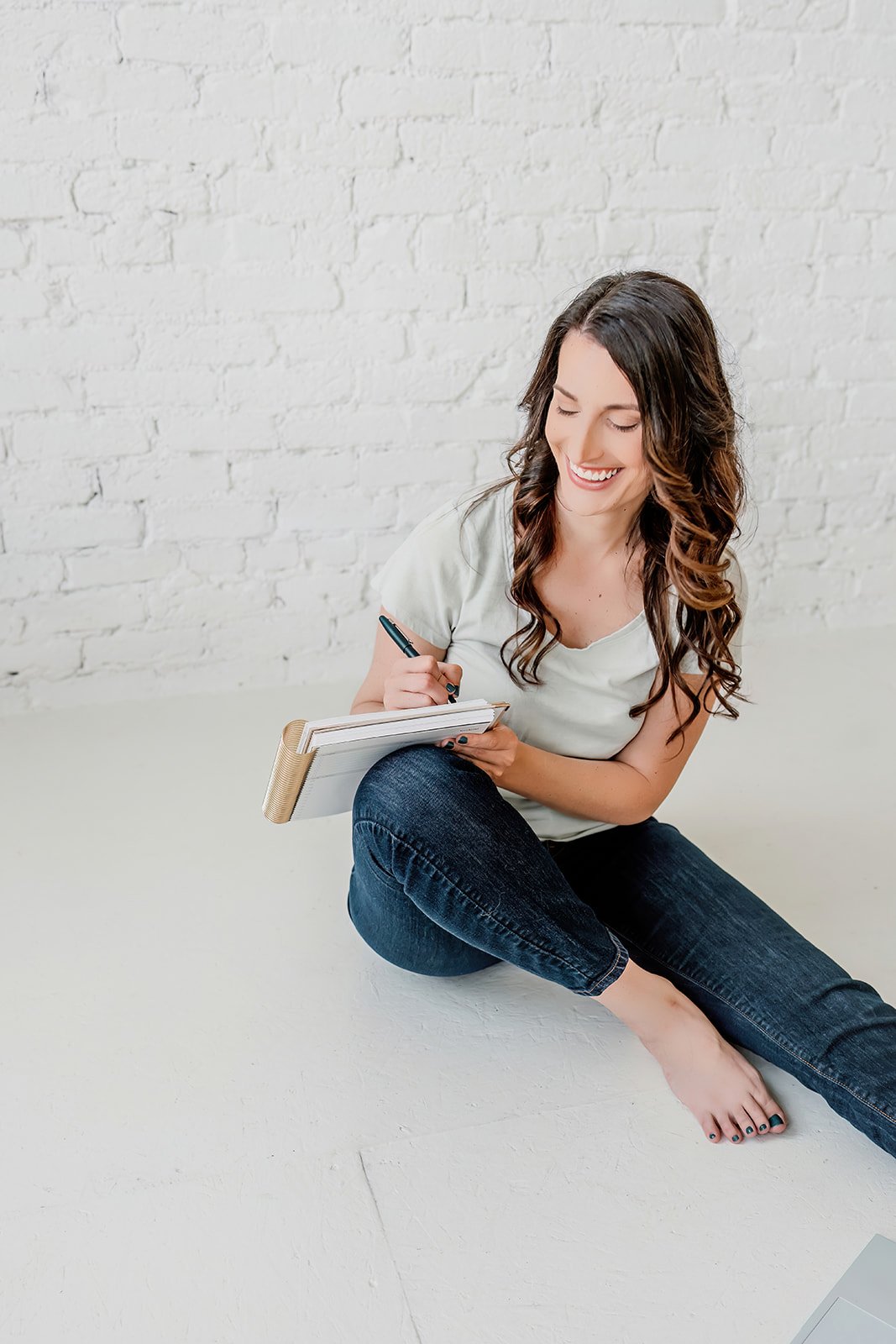 A young woman sitting on the floor near a white brick wall, holding a notebook and writing with a pen, smiling happily.
