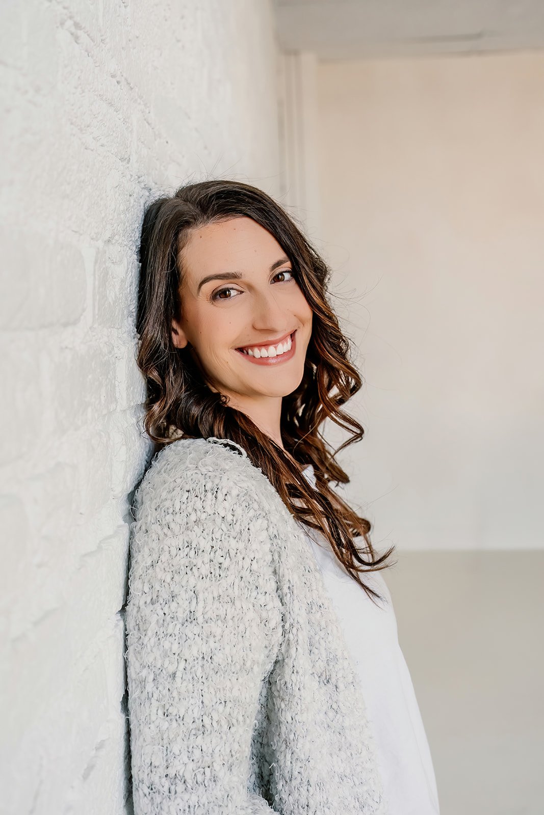 Woman with curly brown hair smiling, leaning against a white brick wall, wearing a white top and light-colored textured cardigan.