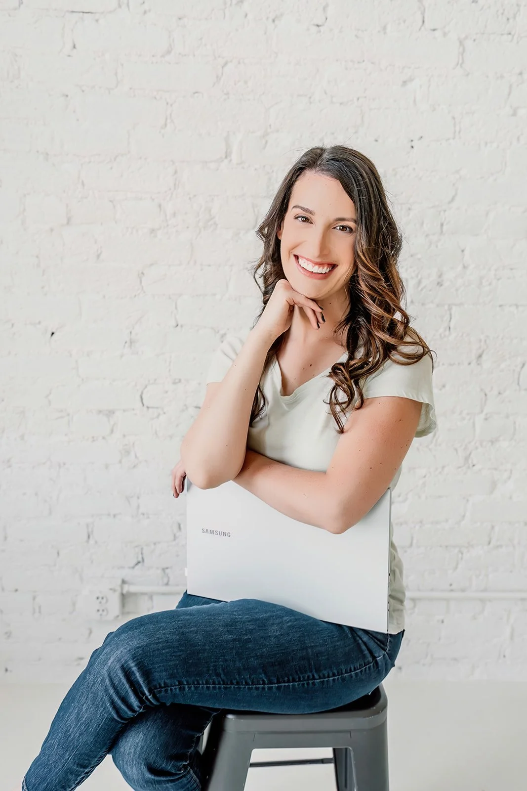 A smiling woman with long brown curly hair sitting on a stool, holding a closed white Samsung laptop on her lap, against a white brick wall background.