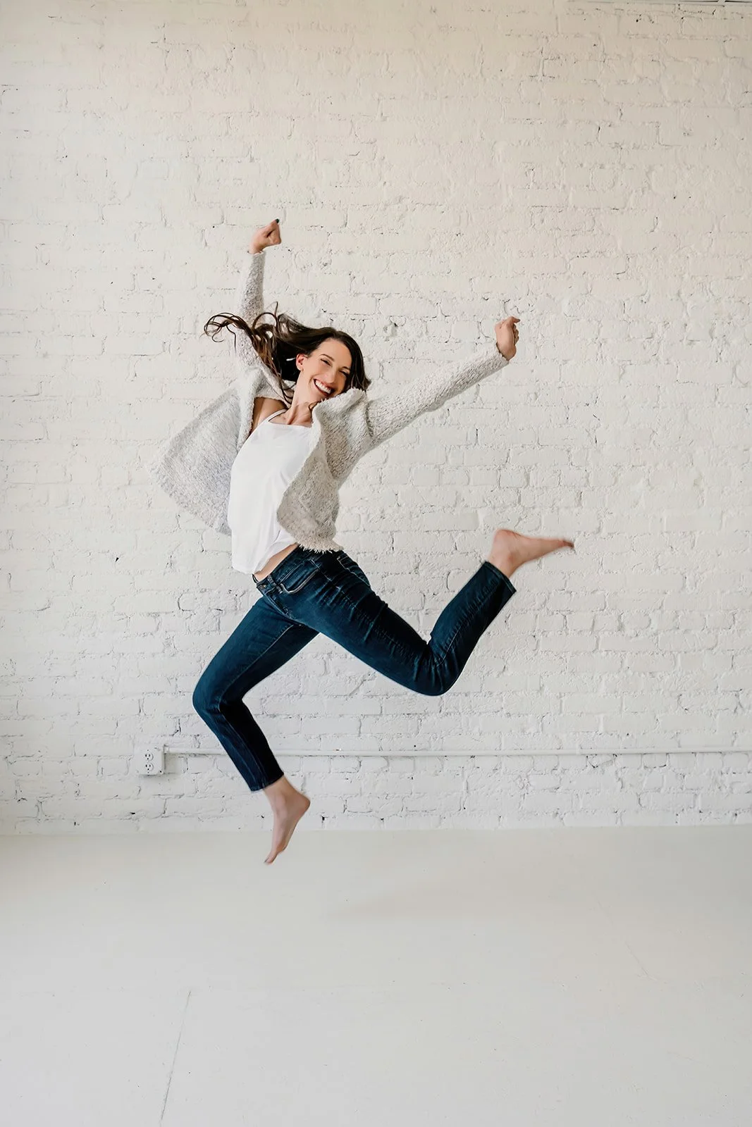A woman jumping in front of a white brick wall, smiling and happy.