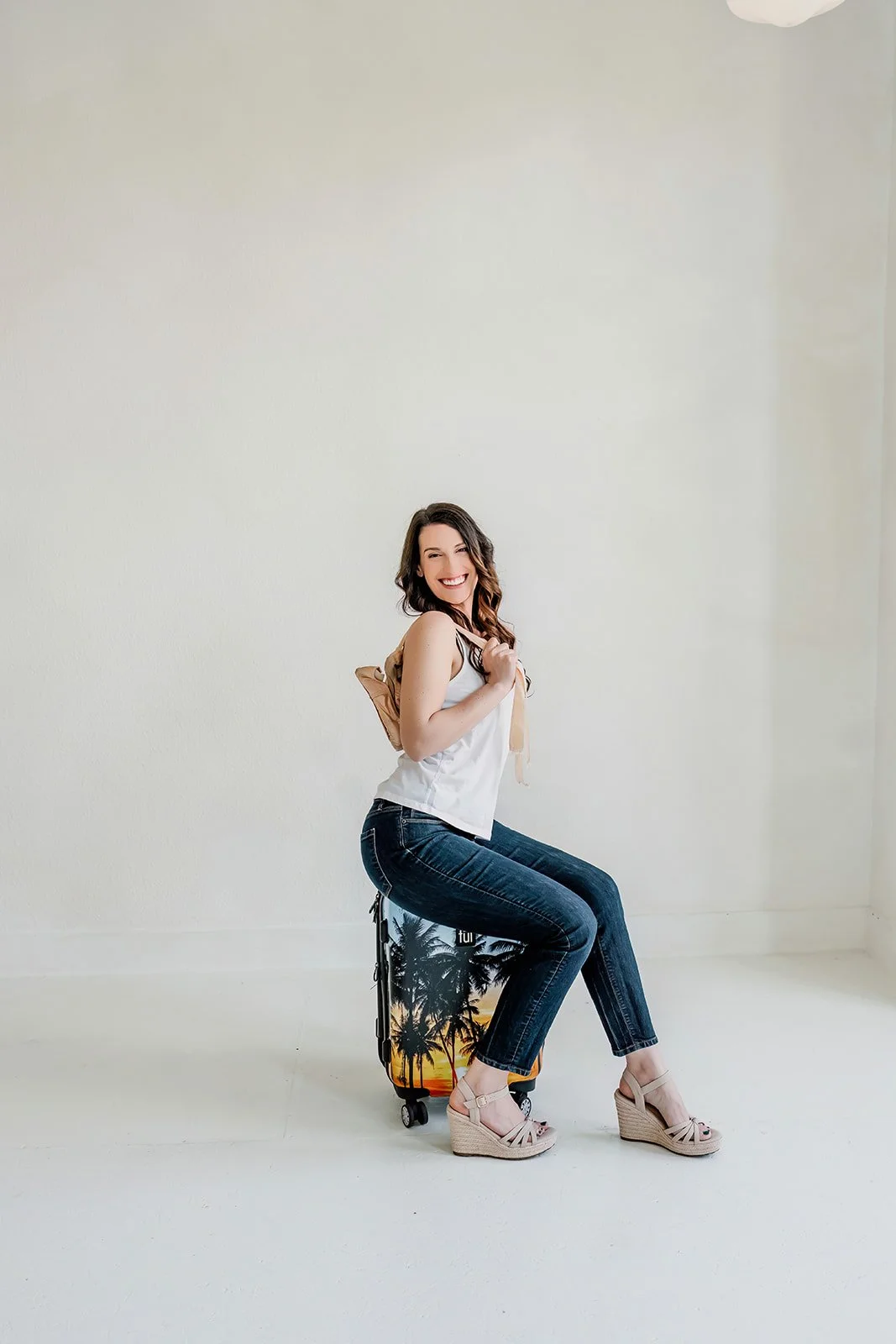 Michelle with long brown hair, smiling and sitting on a small rolling suitcase with a tropical beach scene with palm trees, against a plain white wall, with pointe shoes over her shoulder.