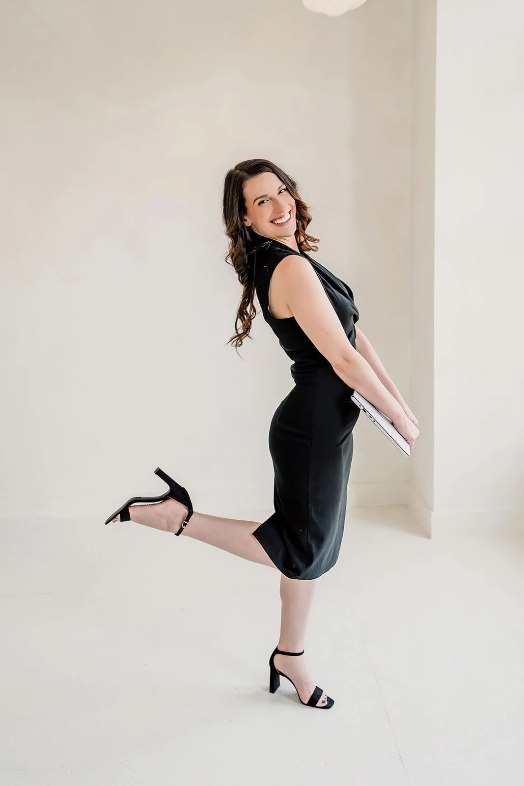 A woman in black high heels and a black dress, standing with one leg raised and bent at the knee, holding a folder and smiling at the camera.
