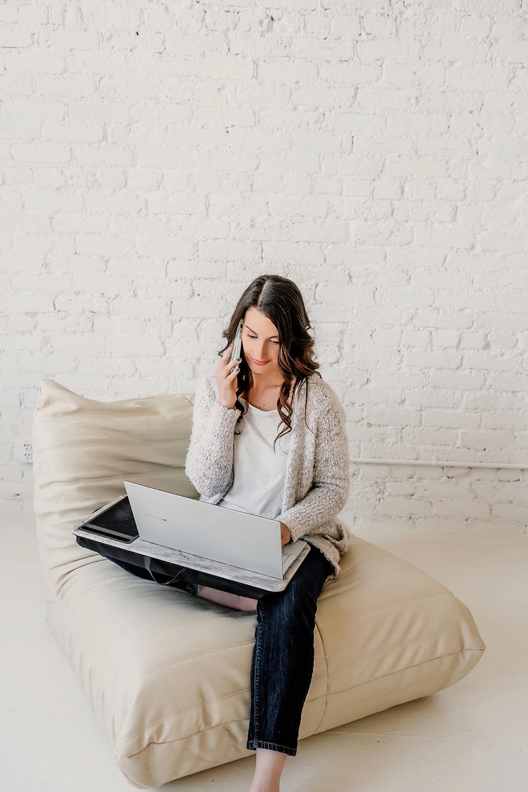 A woman sitting on a beige sofa, talking on a phone, with a laptop on her lap and a smartphone beside her, in front of a white brick wall.