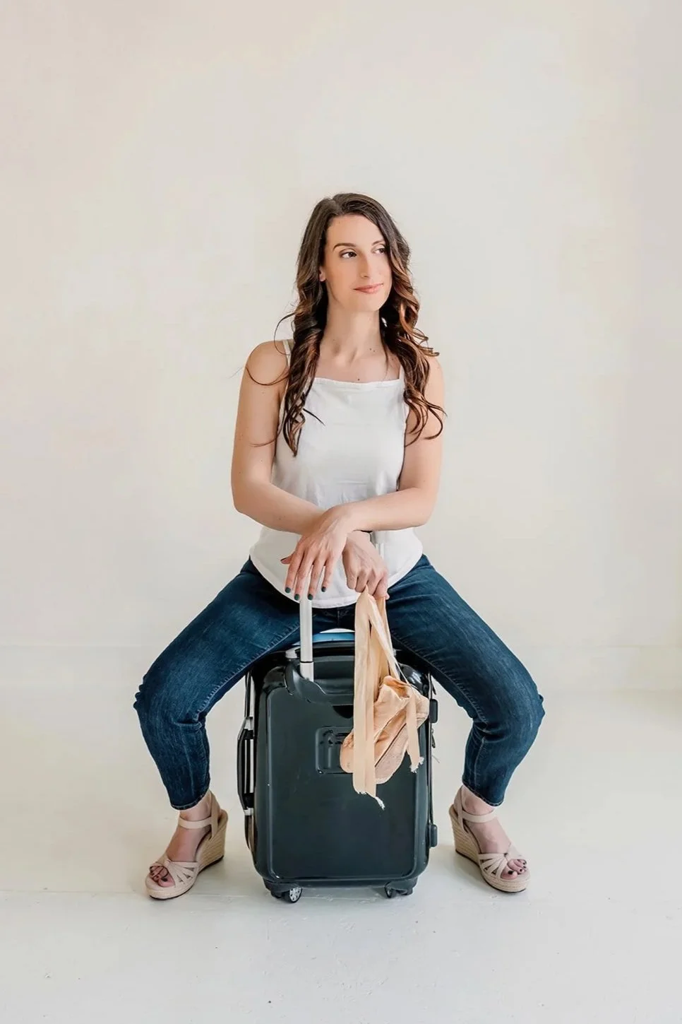 Michelle is sitting on a suitcase with one hand resting on the handle, against a plain white background, wearing a white cami, blue jeans, and wedges, with pointe shoes hanging from her left hand.