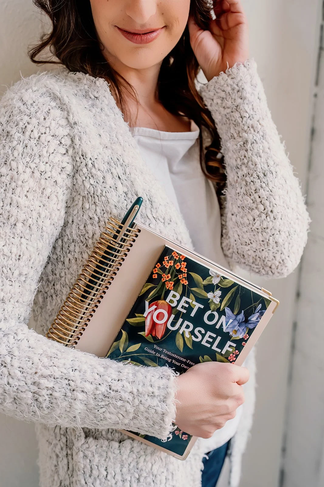 Michelle holding a floral-themed book titled 'Bet On Yourself' by her mentor, Sara Turner, and a beige spiral notebook with a green pen, wearing a white sweater and white shirt.