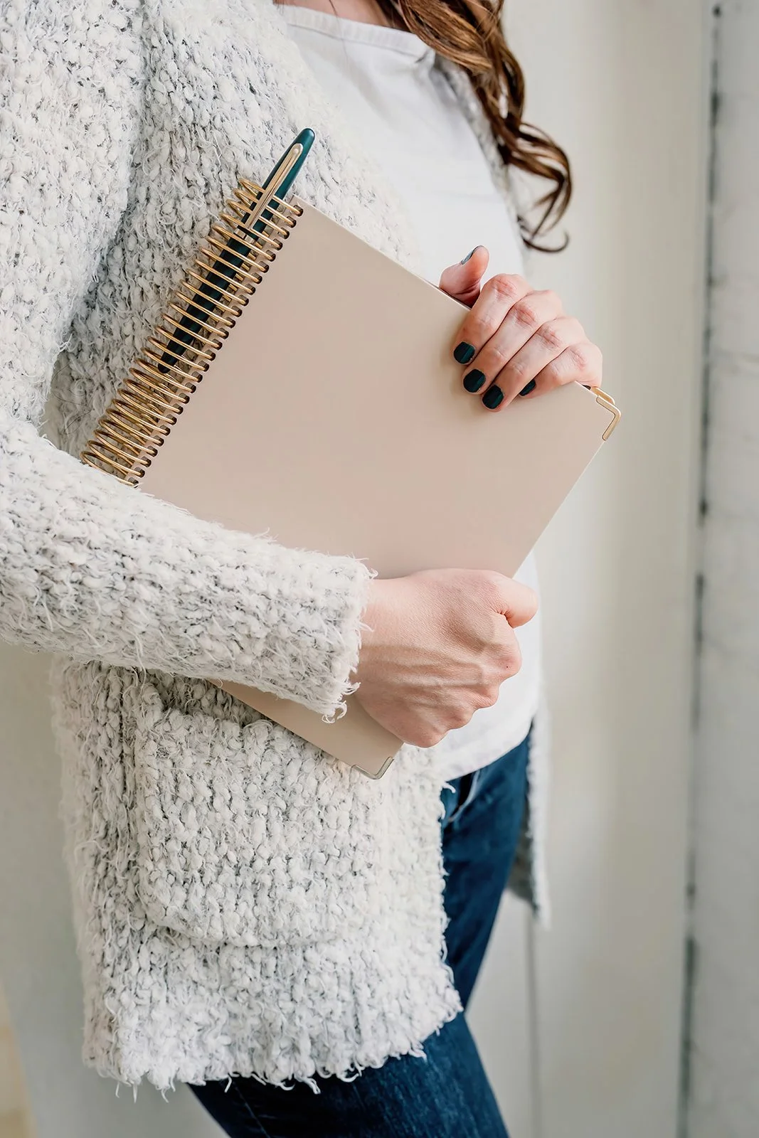 Michelle is holding a notebook and a pen close to her chest, wearing a beige textured sweater and blue jeans, with long wavy hair and neatly painted dark teal nails.