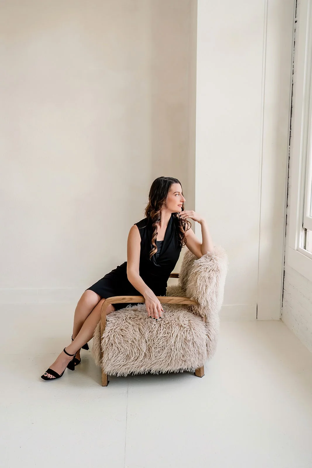 A woman in a black dress sitting on a furry beige armchair near a window in a bright room.