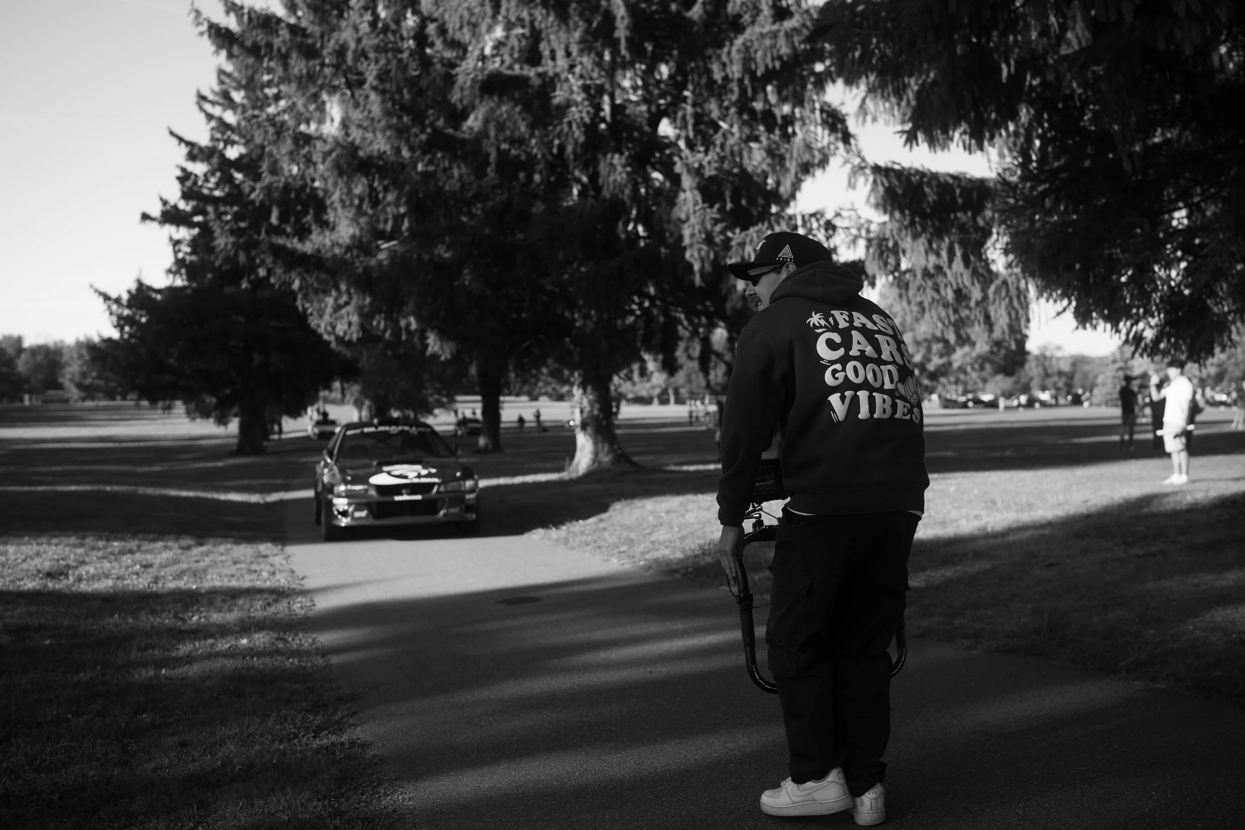 A man walking with a camera rig in a park with large trees, a race car parked on the grass, and a few other people in the background.