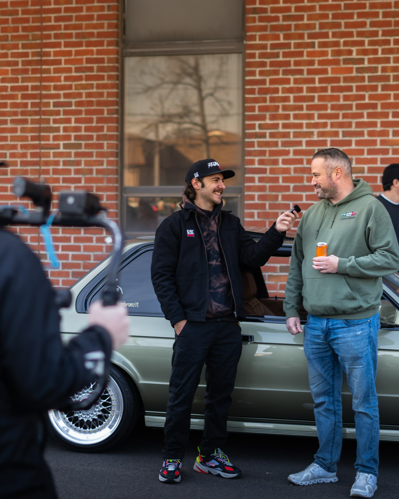 Two men are standing beside a light green car, having a conversation outdoors in front of a brick wall. One man is wearing a black jacket, black cap, and colorful sneakers, holding a microphone. The other man is wearing a green hoodie and jeans, and holding a can of soda.