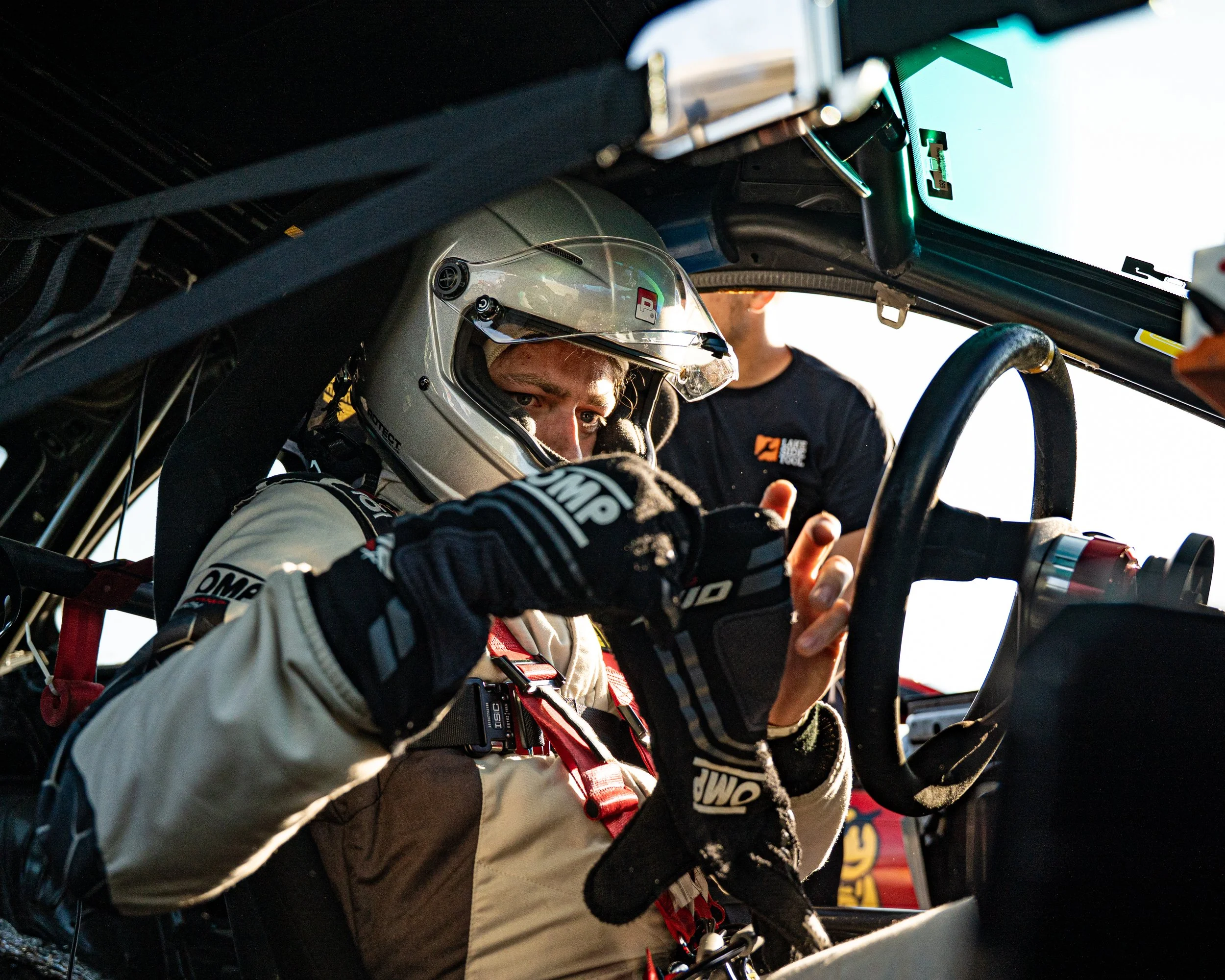 Race car driver wearing a helmet and racing suit inside a race car, with a team member near the window.