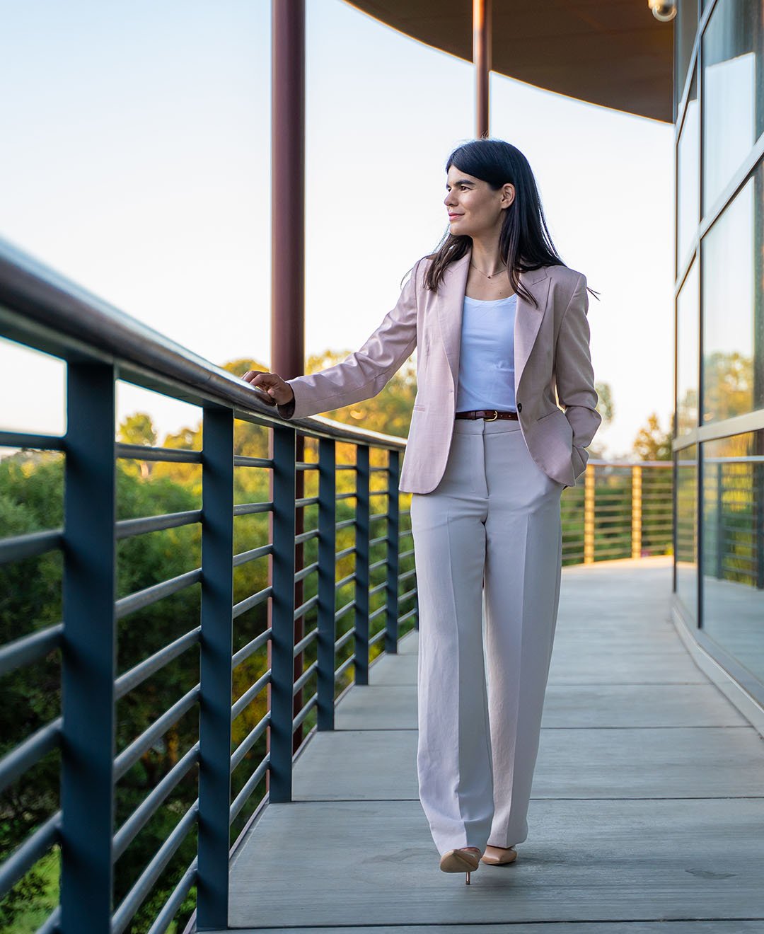 A woman in a light pink blazer and white pants walking on a balcony with a railing, with trees and a sunset sky in the background.
