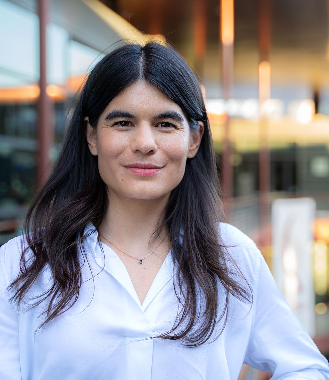 A woman with long dark hair wearing a white blouse and a necklace, smiling outdoors with a modern building in the background.