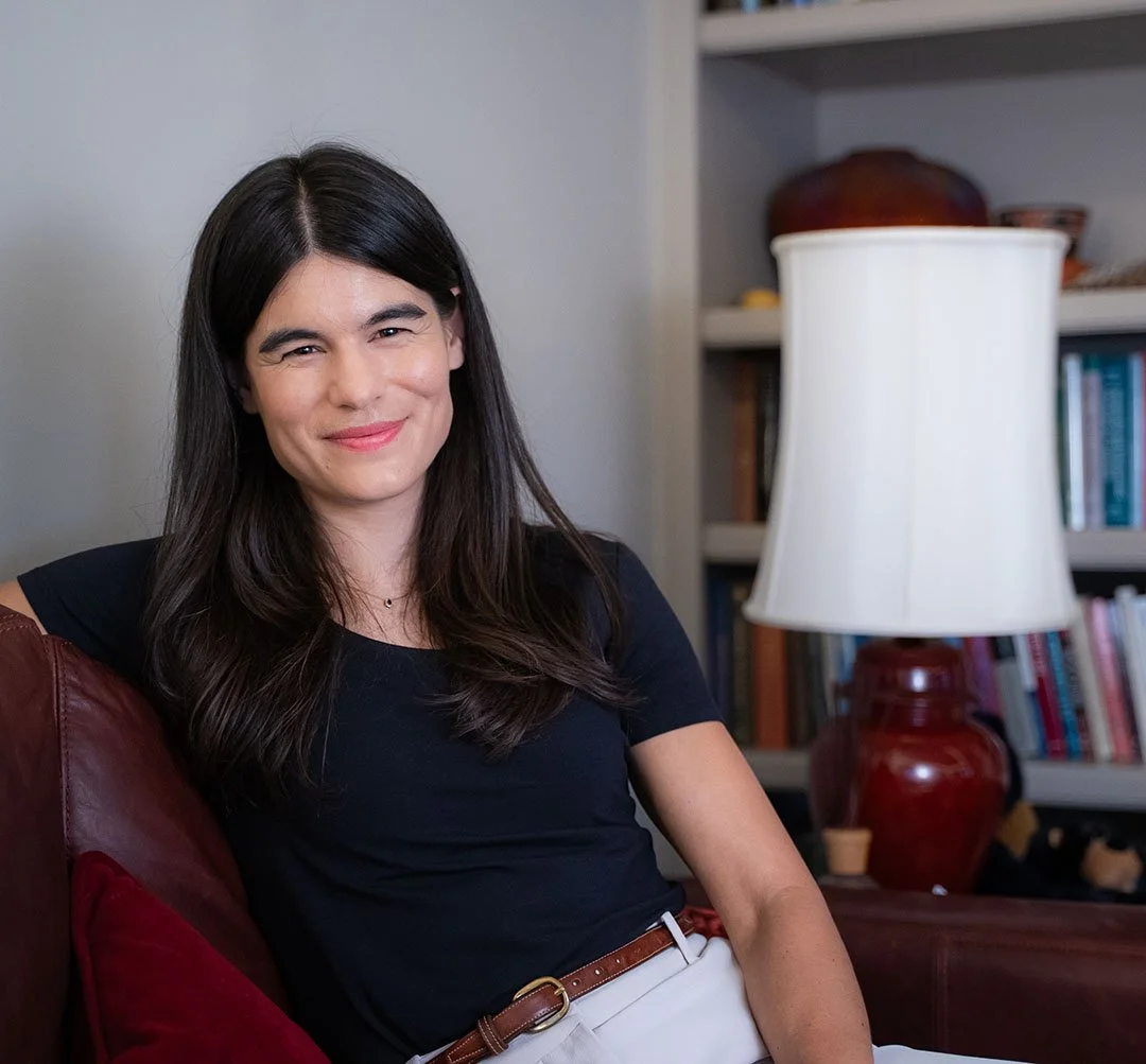 A woman with long dark hair smiling and sitting on a leather couch, with a bookshelf and a large white lampshade behind her.