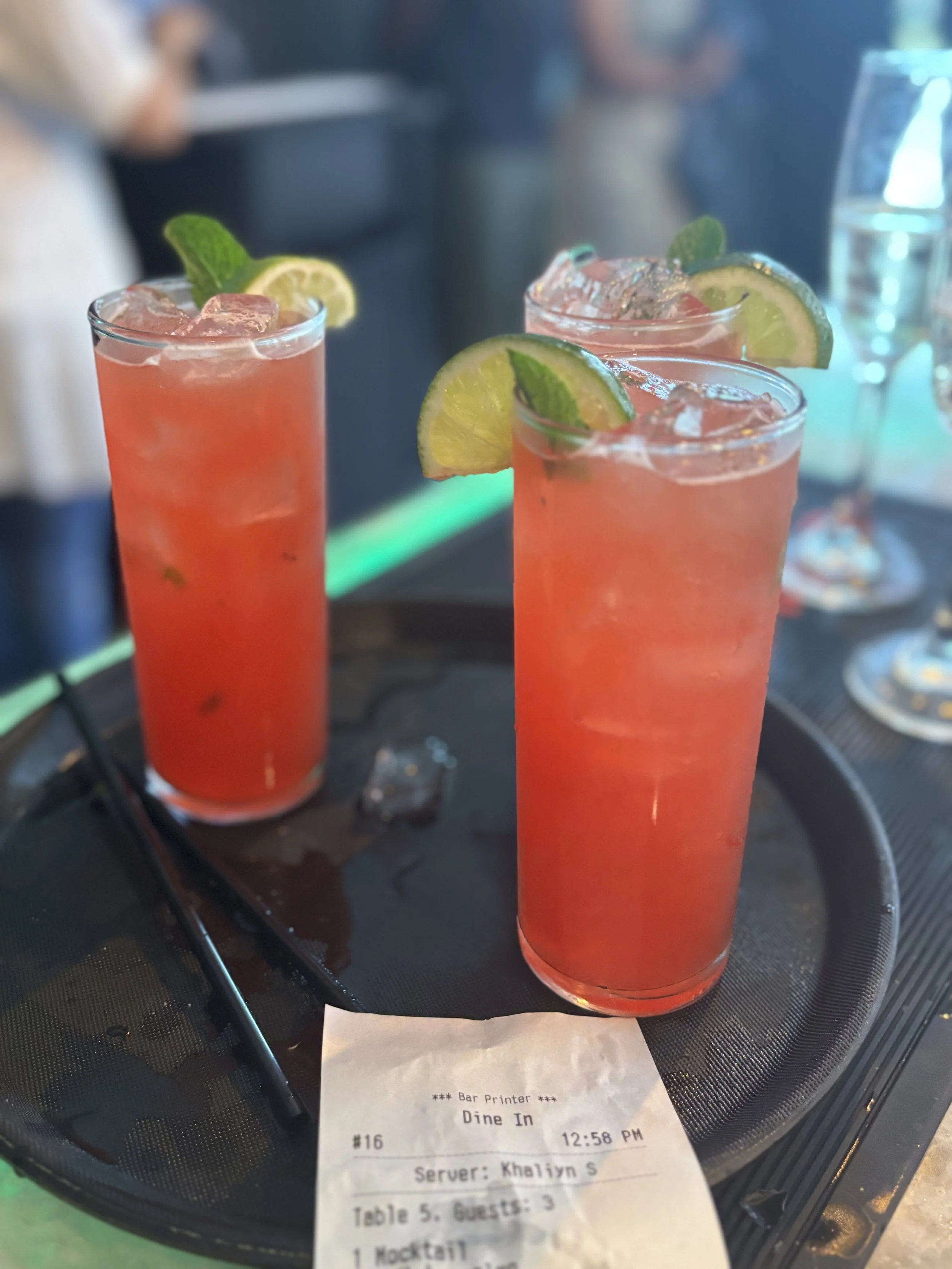 Three tall glasses of pink cocktails garnished with lime slices and mint leaves on a tray, with a receipt beneath them.