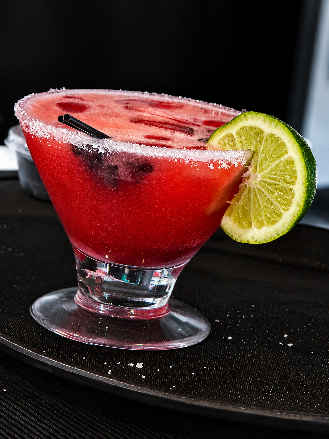 A pink watermelon cocktail garnished with a lime wedge, served in a glass with a salted rim and two black straws.