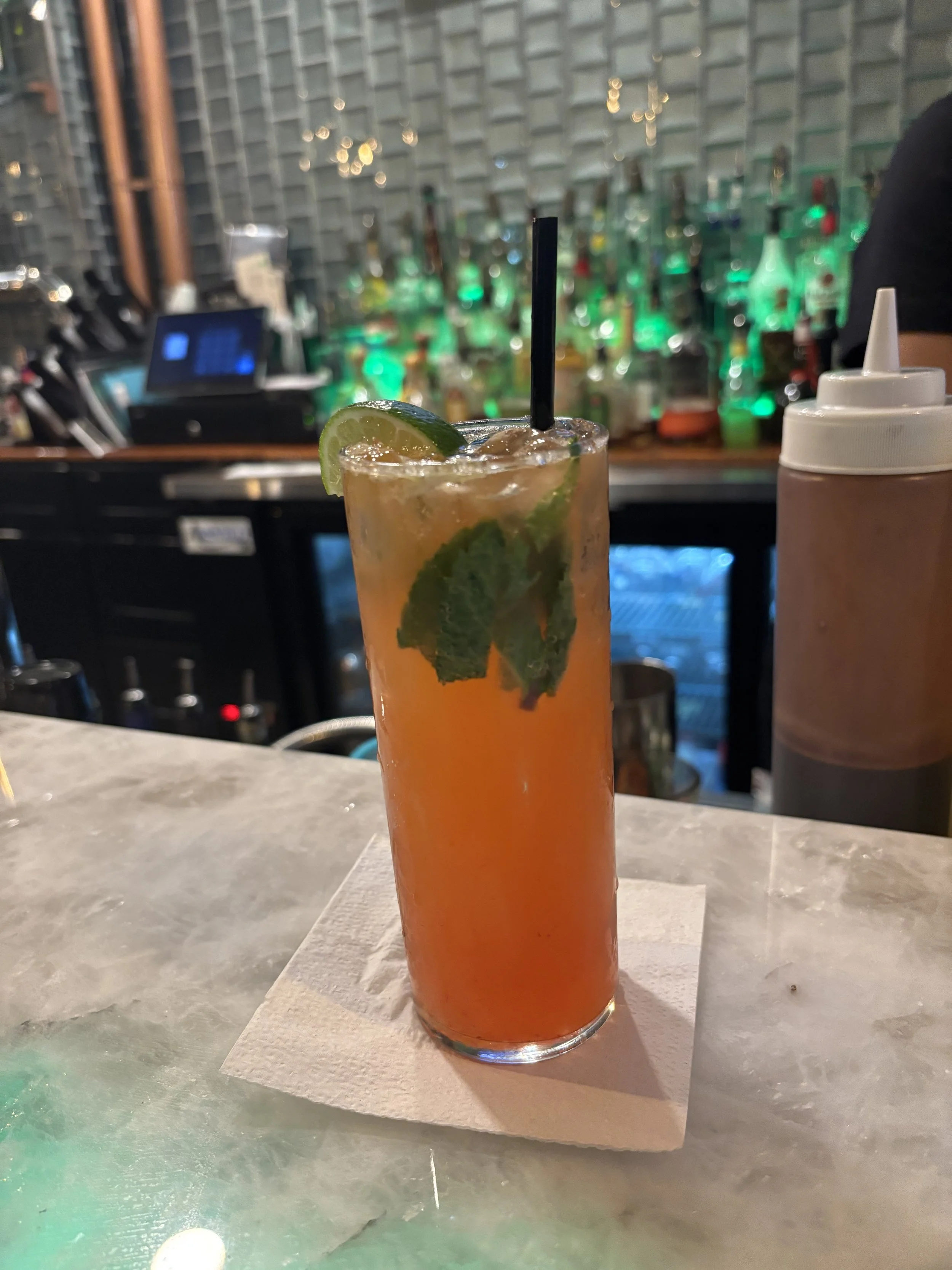 A tall glass of orange-colored cocktail with lime wedge and mint leaves, on a napkin at a bar counter. The blurred background shows bar shelves with bottles and bar equipment.