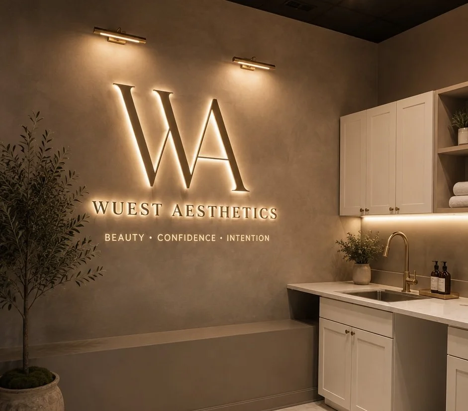 Interior of a wellness or aesthetic clinic with illuminated sign reading 'Wuest Aesthetics' and the words 'Beauty, Confidence, Intention' underneath. The space features a kitchen-like area with white cabinets, a gold faucet, and decorative plants.