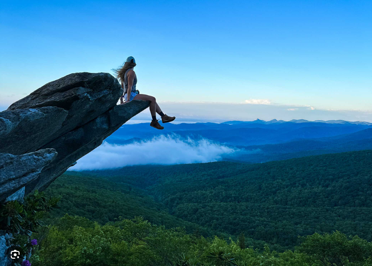 A person sitting on the edge of a large rock over a mountain landscape with clouds and forest below.