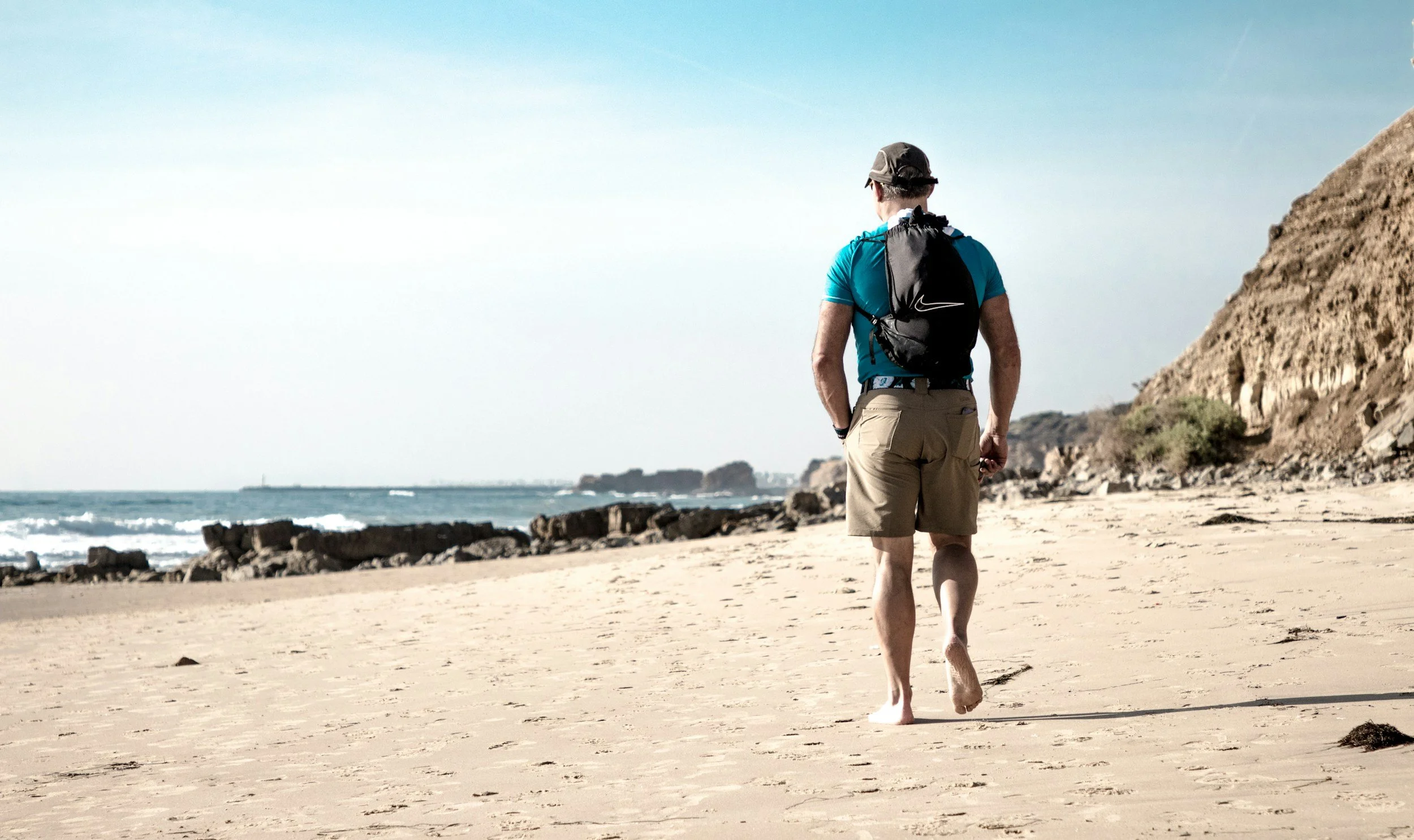 A man walking alone on a sandy beach with rocks, ocean waves, cliffs, and a clear sky in the background.