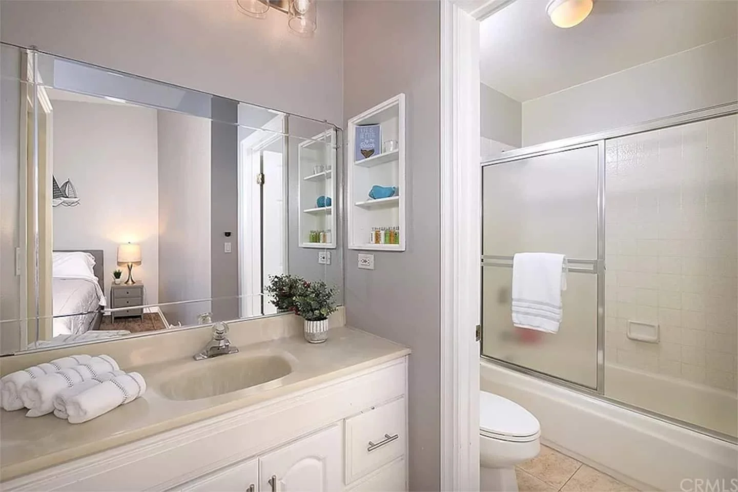 Bathroom with a beige vanity and sink, a large mirror, a small plant, and towels; a toilet; a glass shower enclosure with a towel hanging on the door; bedroom visible through the mirror reflection.